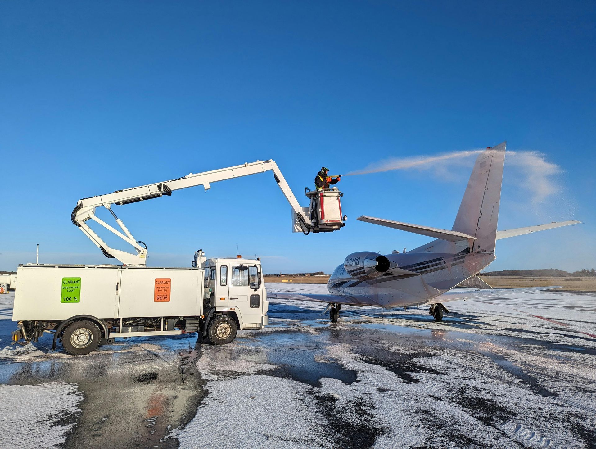 Truck with lift spraying de-icing fluid on a parked private jet at an airport on a snowy day.