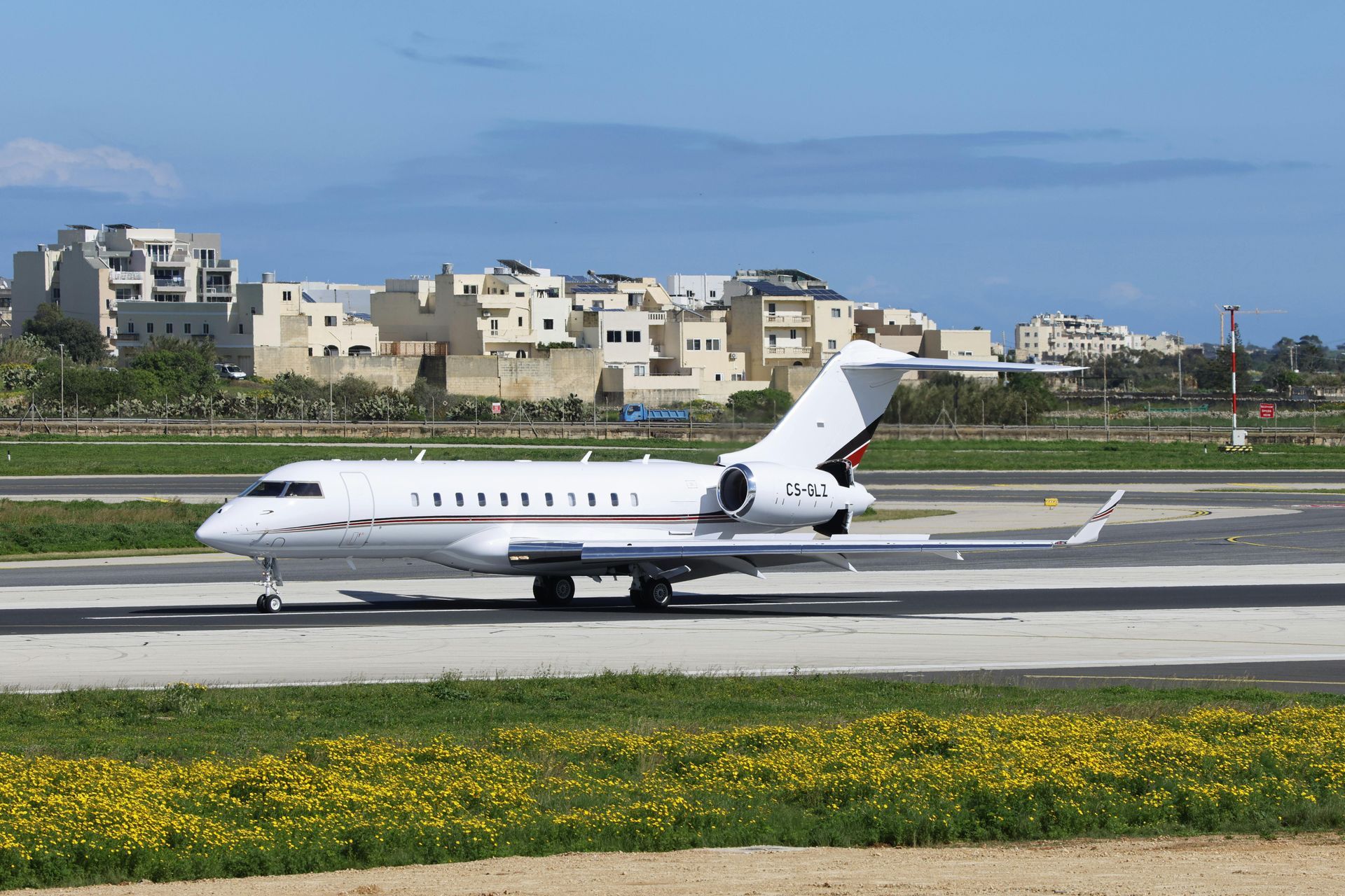 White private jet on a runway, buildings in the background, blue sky.