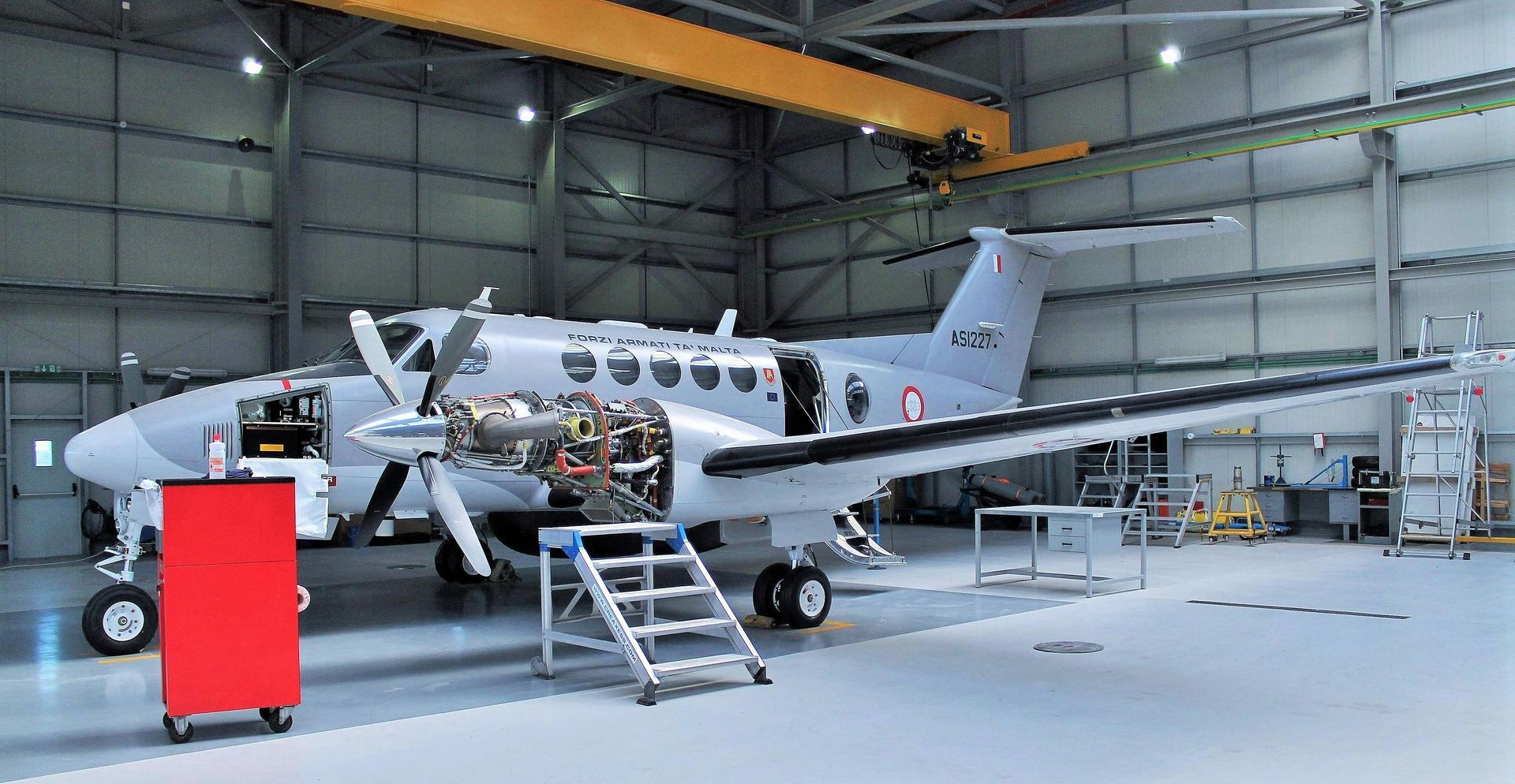 Aircraft in a hangar, undergoing maintenance with open engine bay. Gray and red hues.