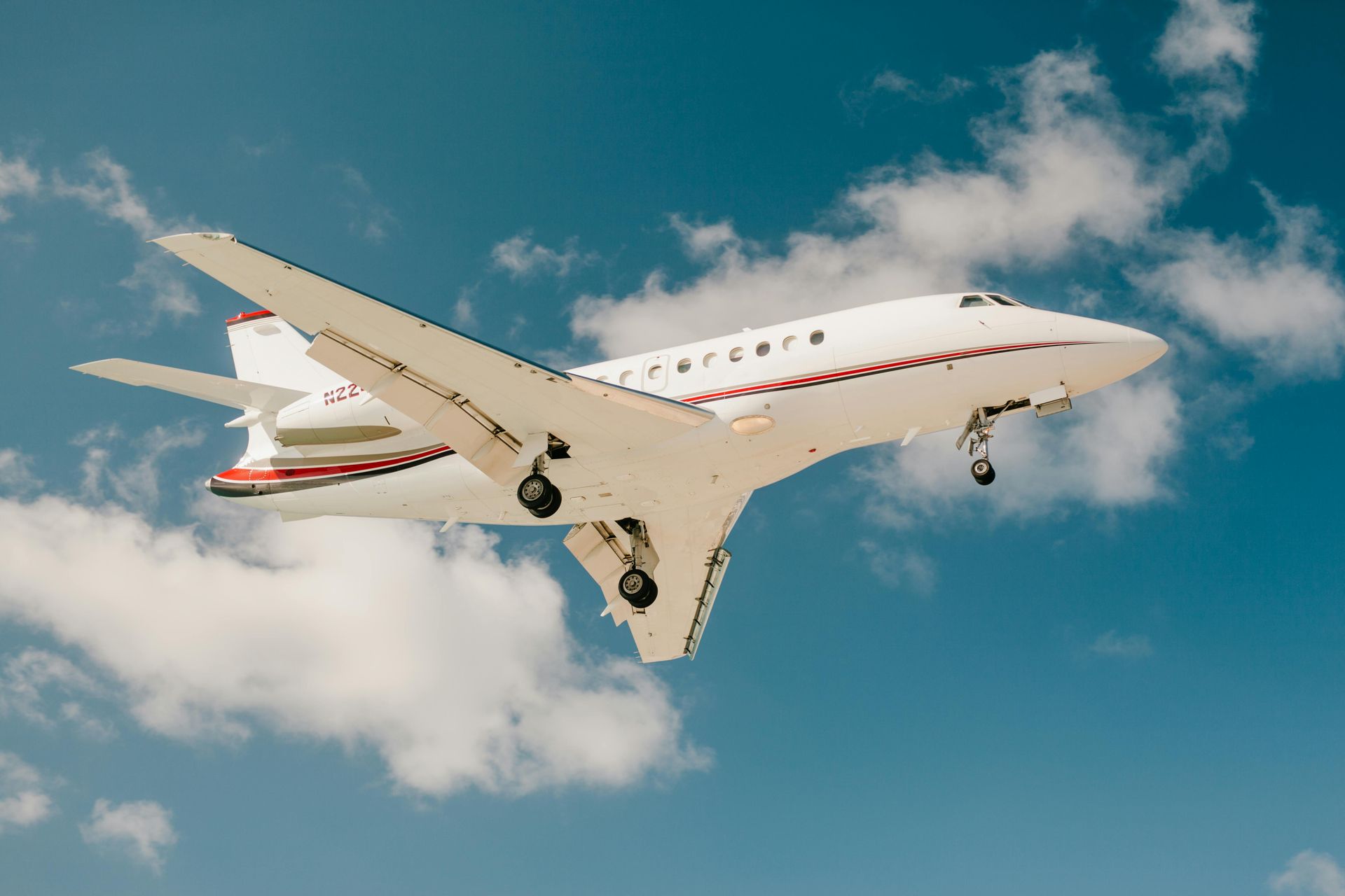 White private jet with red accents against a blue sky with clouds, landing gear down.
