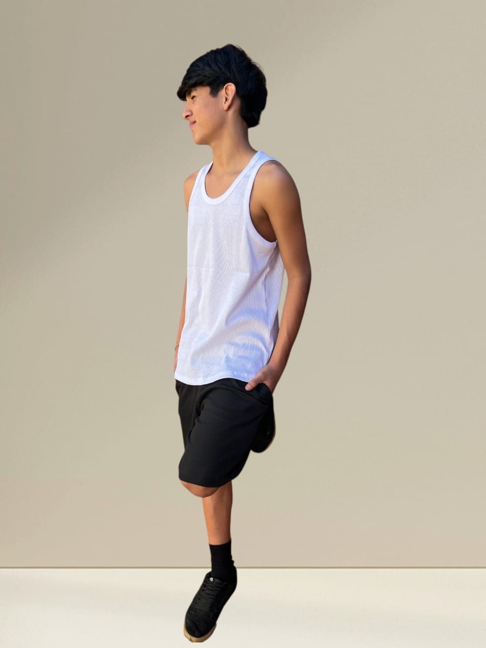 Persona con camiseta blanca de tirantes y pantalones cortos negros con las manos en los bolsillos; de pie sobre una pierna.