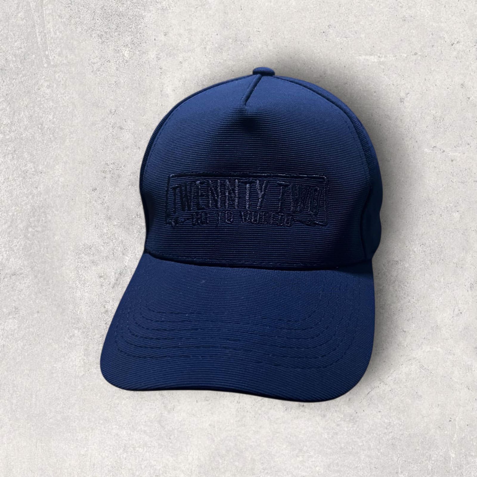 Gorra de béisbol azul marino con un logotipo rectangular en relieve de color azul oscuro en la parte delantera.