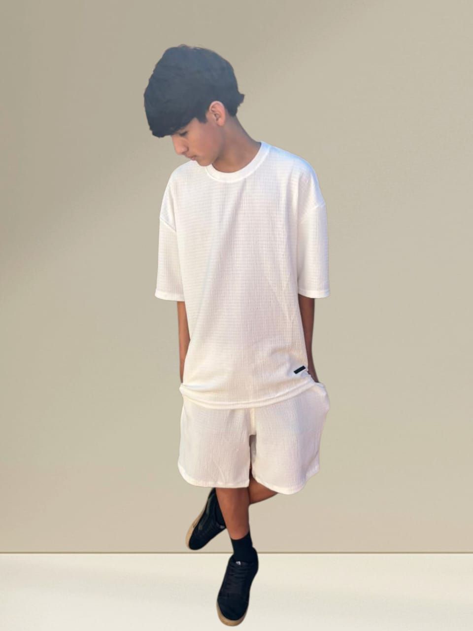 Persona con camiseta blanca, pantalones cortos, zapatos negros y gorro de lana, mirando hacia abajo. Fondo neutro.