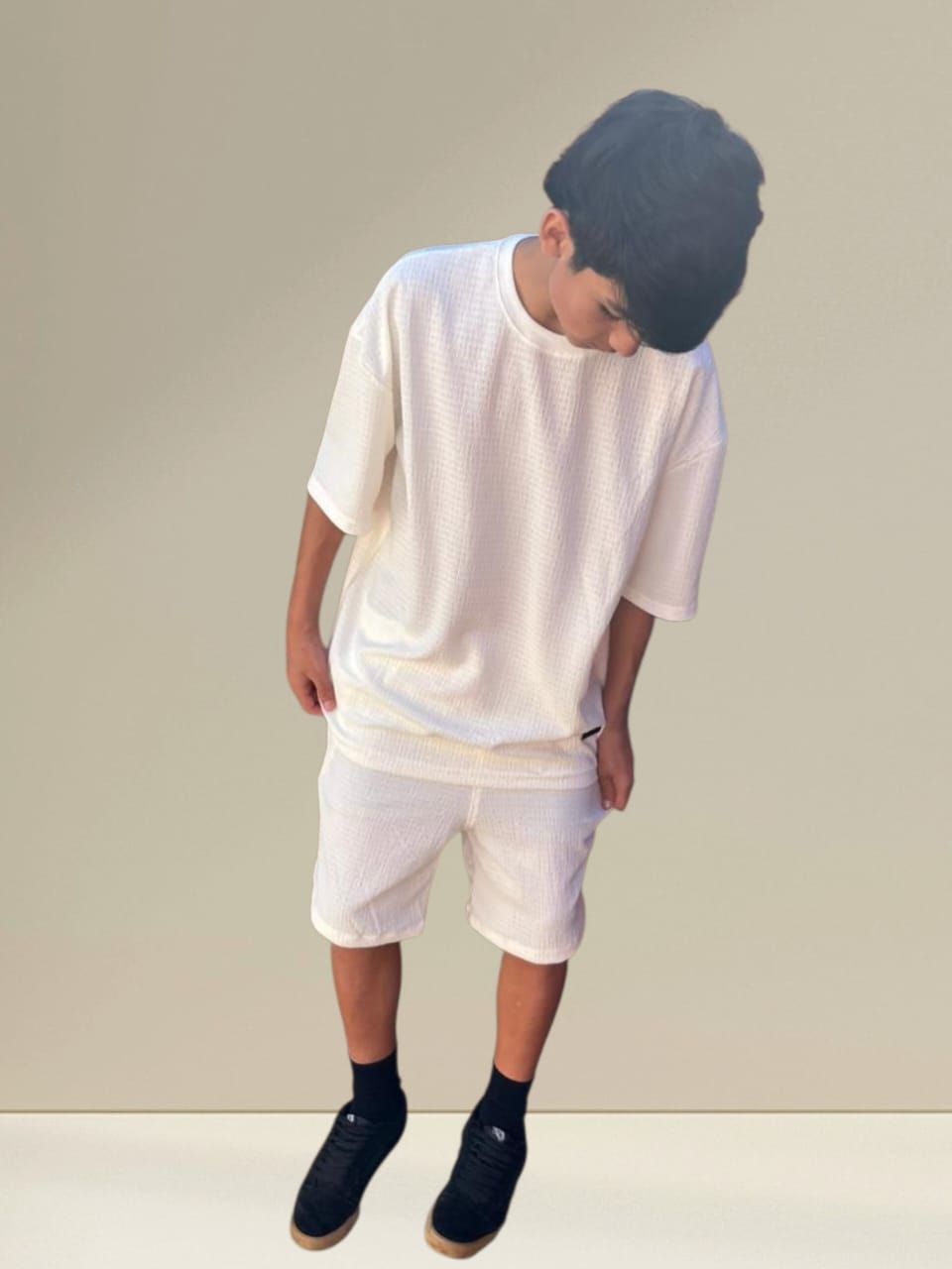 Persona con camiseta blanca, pantalones cortos, zapatos negros y gorro de lana, mirando hacia abajo. Fondo neutro.