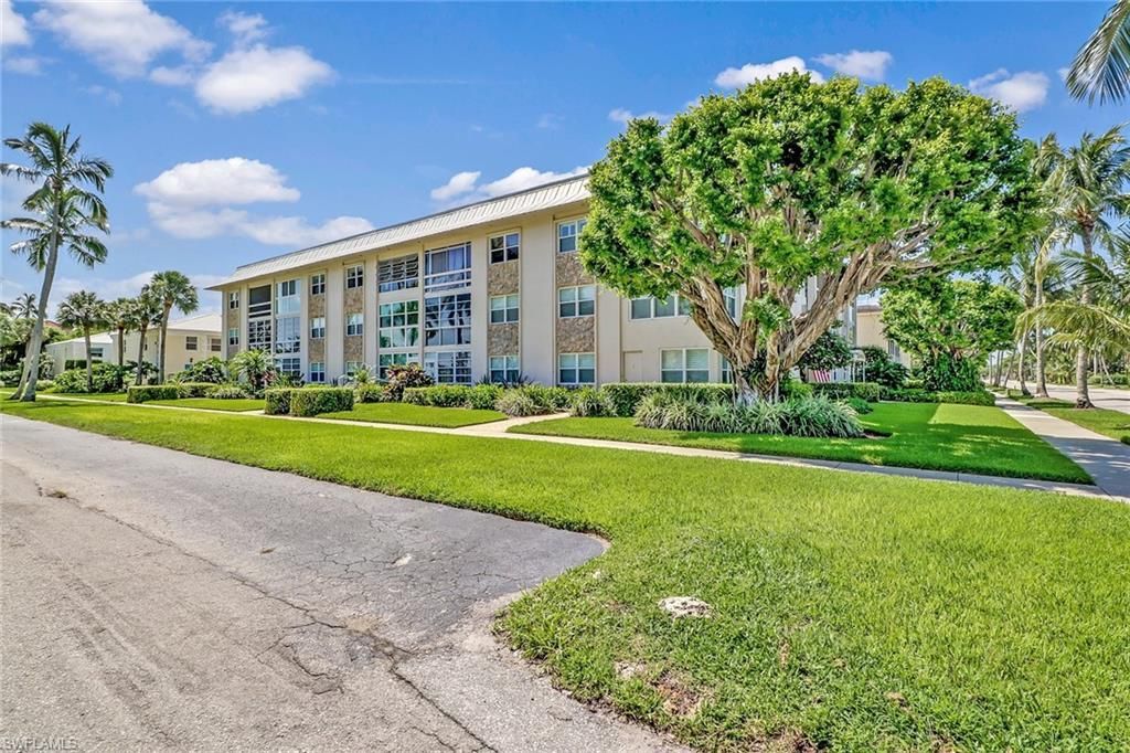 Three-story apartment building with palm trees, green lawn, and a paved road under a blue sky.