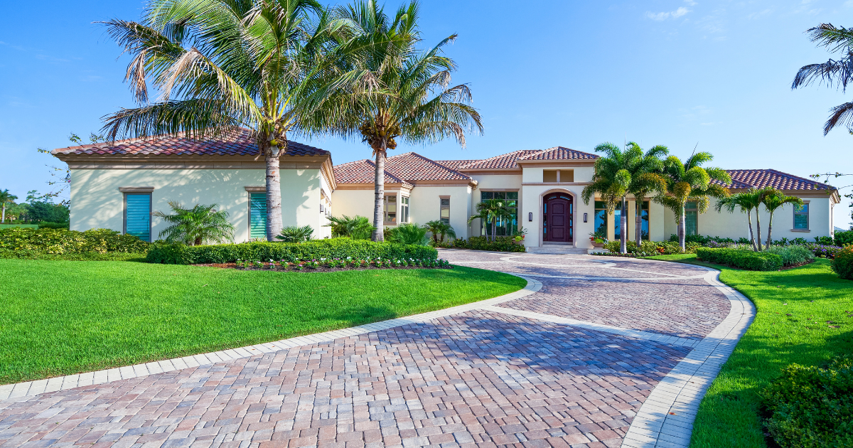 Single-story stucco house with a curved brick driveway, palm trees, and a lush green lawn under a bright sky