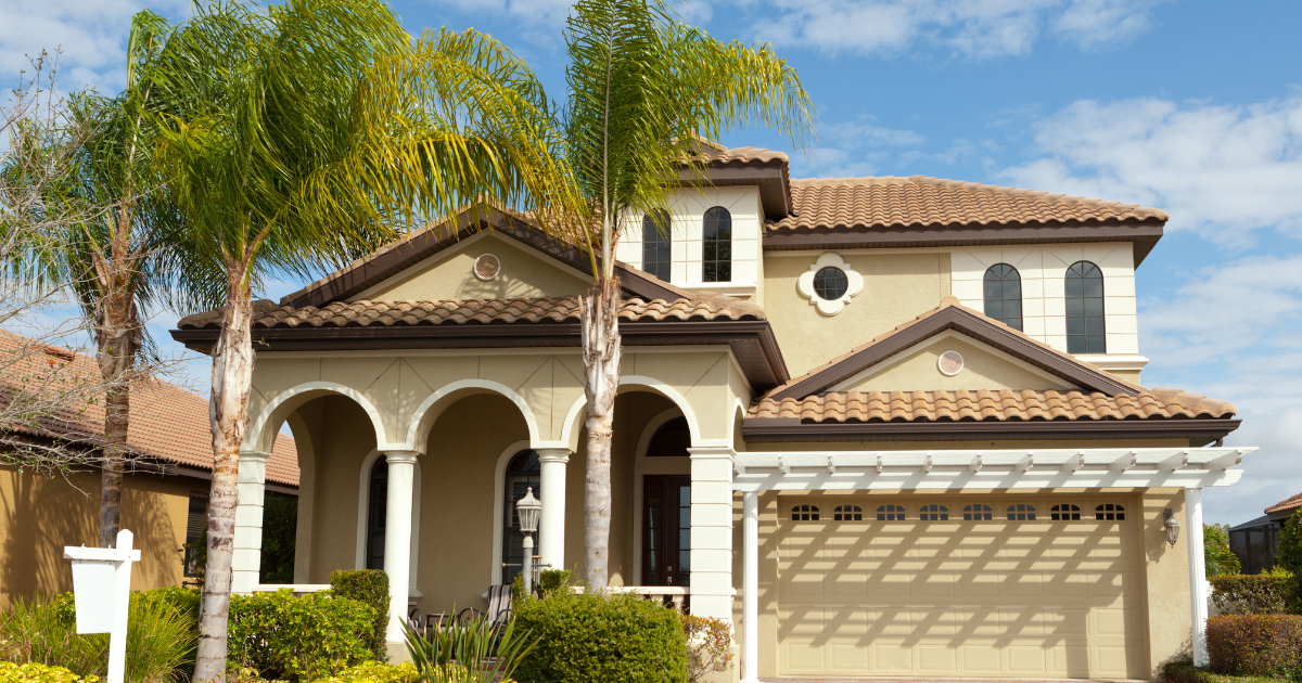 Two-story beige Mediterranean-style house with a tiled roof, arched porch, and palm trees in front