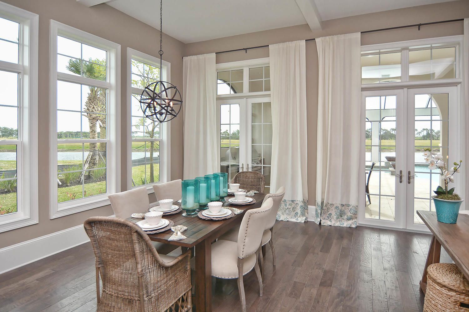 Bright dining room with large windows, wooden table, wicker chairs, and teal centerpieces.