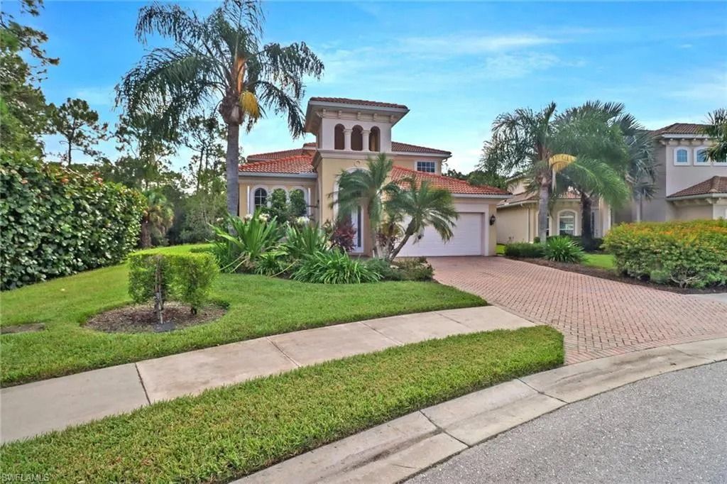 Mediterranean-style house with a brick driveway, palm trees, and manicured front yard under a blue sky