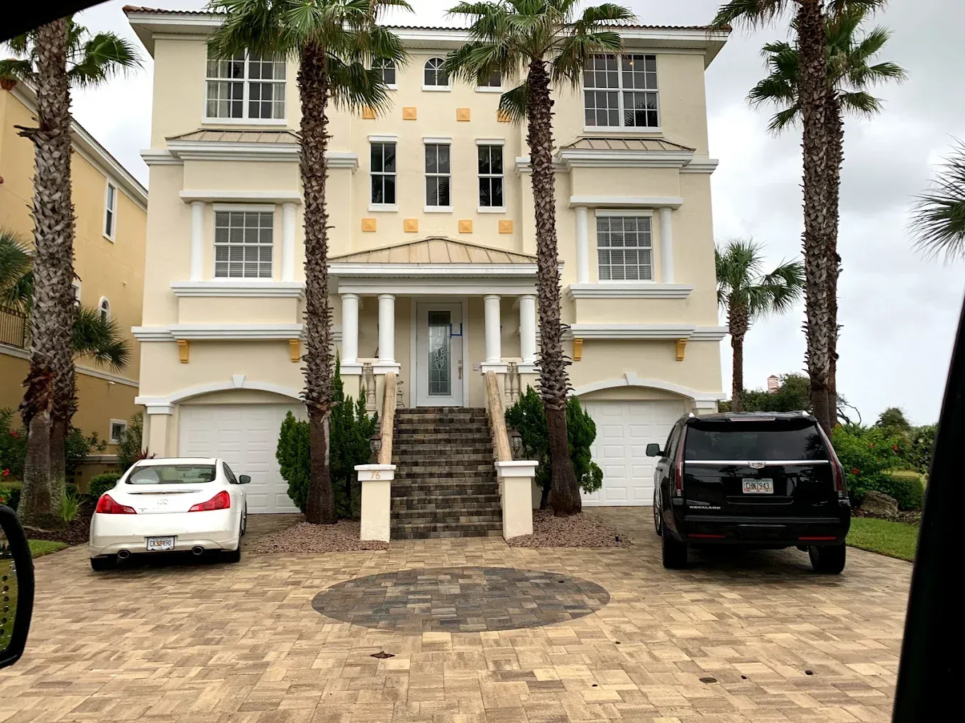 Two cars are parked in front of a large house with palm trees.