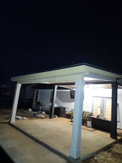 Nighttime view of a concrete patio with a white-columned awning. A trailer is visible in the background.