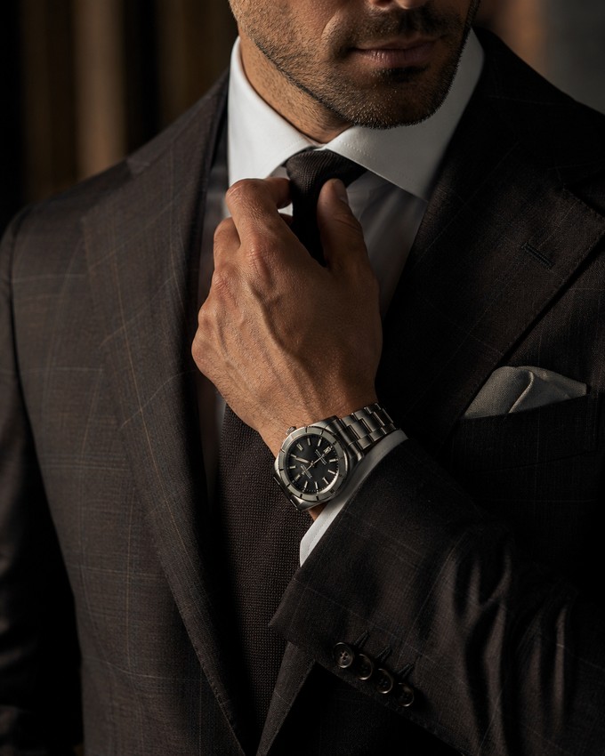 Uomo che si sistema una cravatta scura con un orologio d'argento, indossa un abito color antracite e un fazzoletto da taschino.