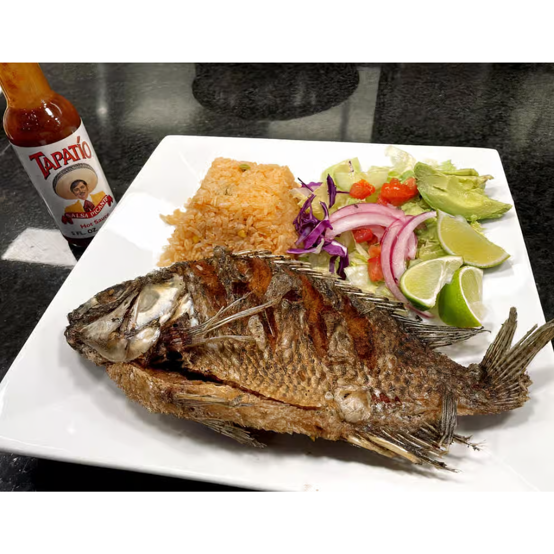 A white plate topped with fried fish , rice and vegetables next to a bottle of tapatio sauce.