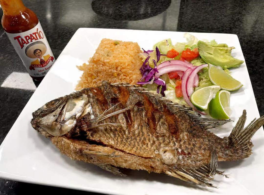 A white plate topped with fried fish rice and vegetables next to a bottle of don julio hot sauce.