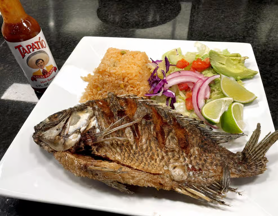 A white plate topped with fried fish rice and vegetables next to a bottle of tabasco sauce.