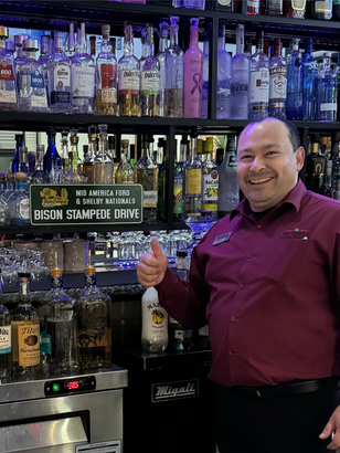 A man in a red shirt is standing in front of a bar giving a thumbs up.