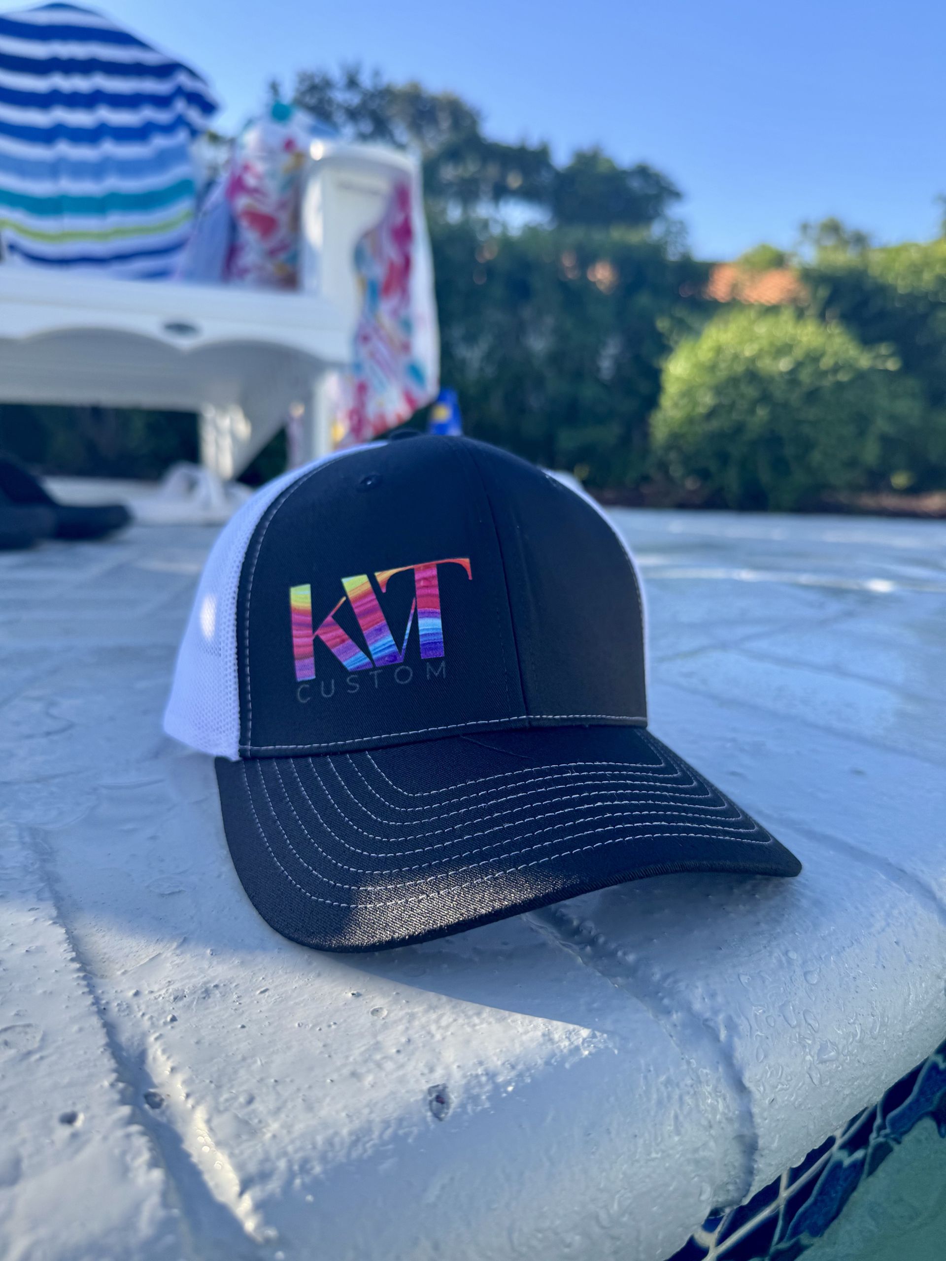 Black and white KT logo trucker hat by a pool with blue water on a sunny day.