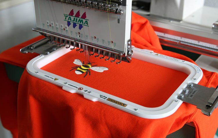 Embroidery machine stitching a bee design onto an orange shirt. White hoop holds the fabric.