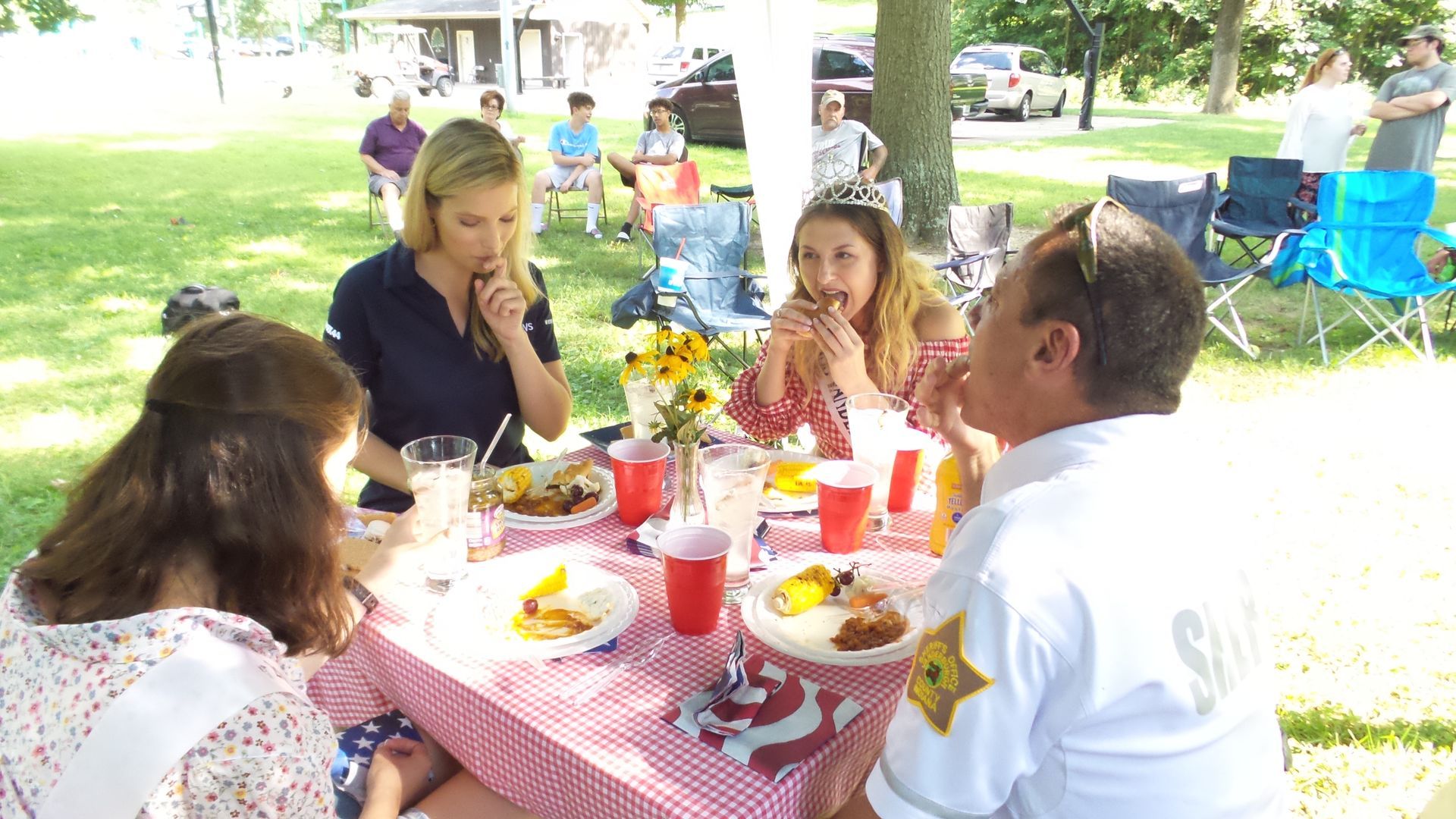 People eating picnic lunch at a park table under trees, with red cups and plates on a red-checkered cloth.