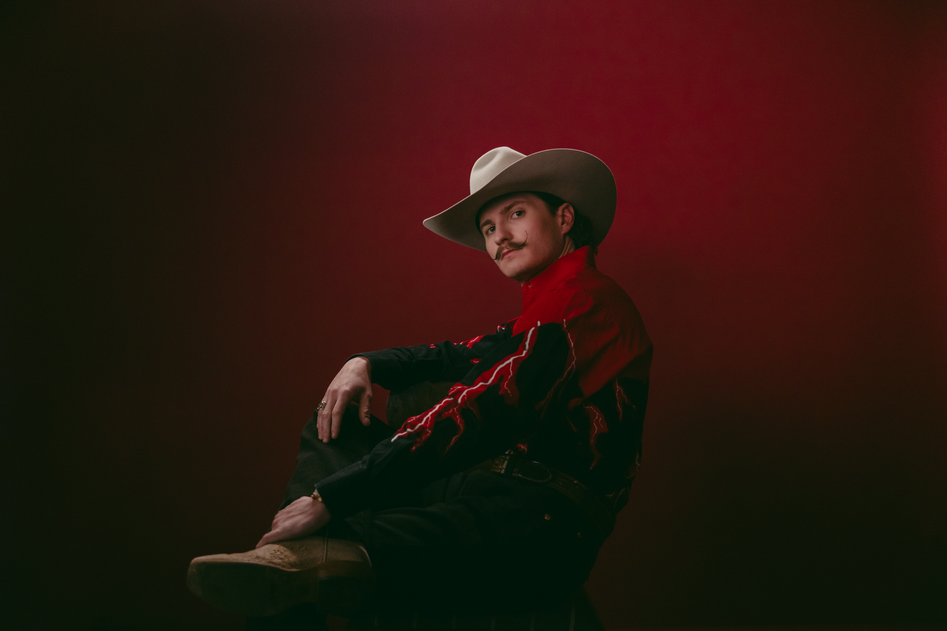 Man in a straw cowboy hat and orange jacket sits cross-legged against a dark background.