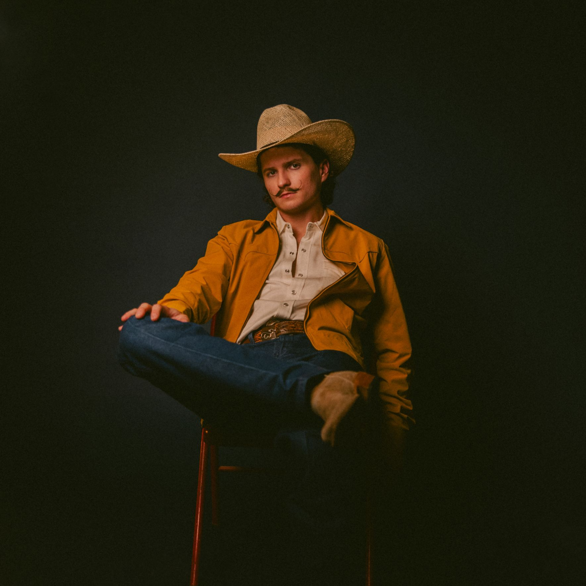 Man in a straw cowboy hat and orange jacket sits cross-legged against a dark background.