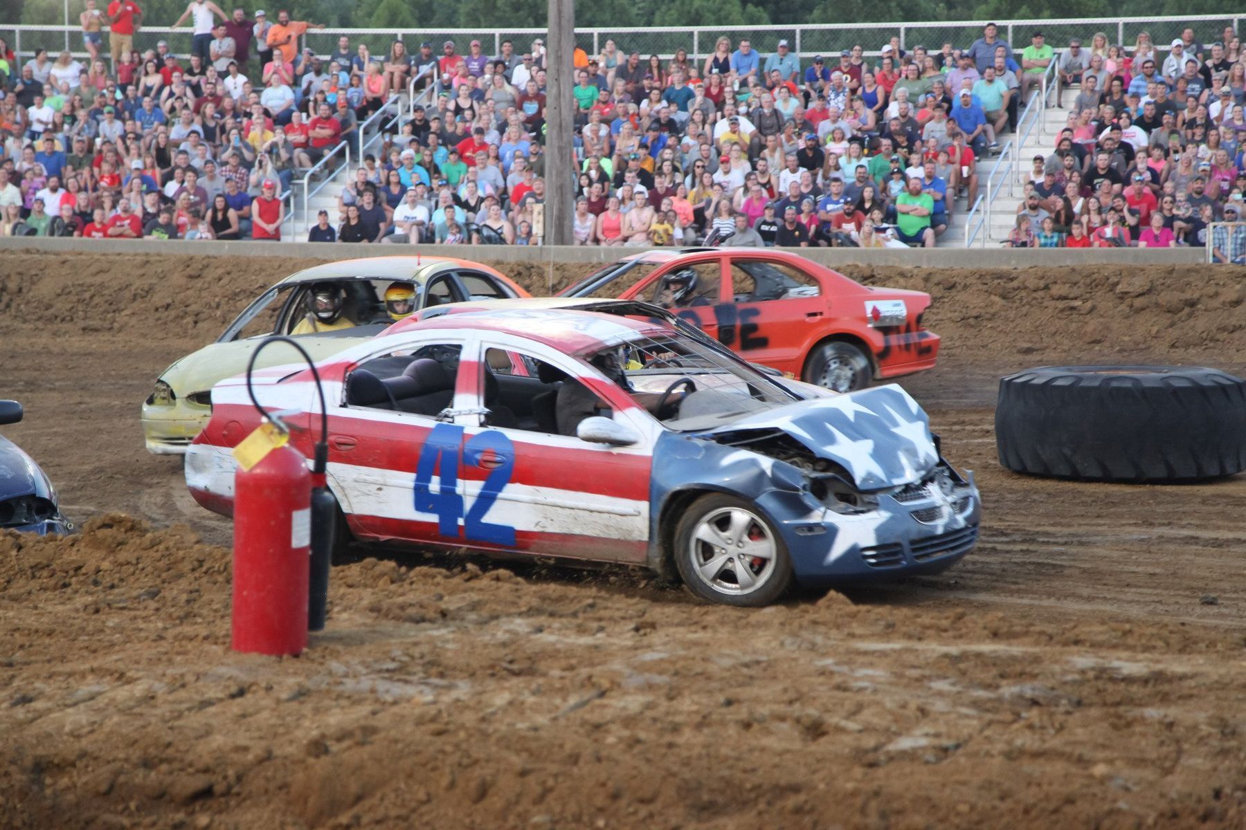 Two stock cars racing on a muddy track, with a red-white-blue car in front and a crowd in the stands.