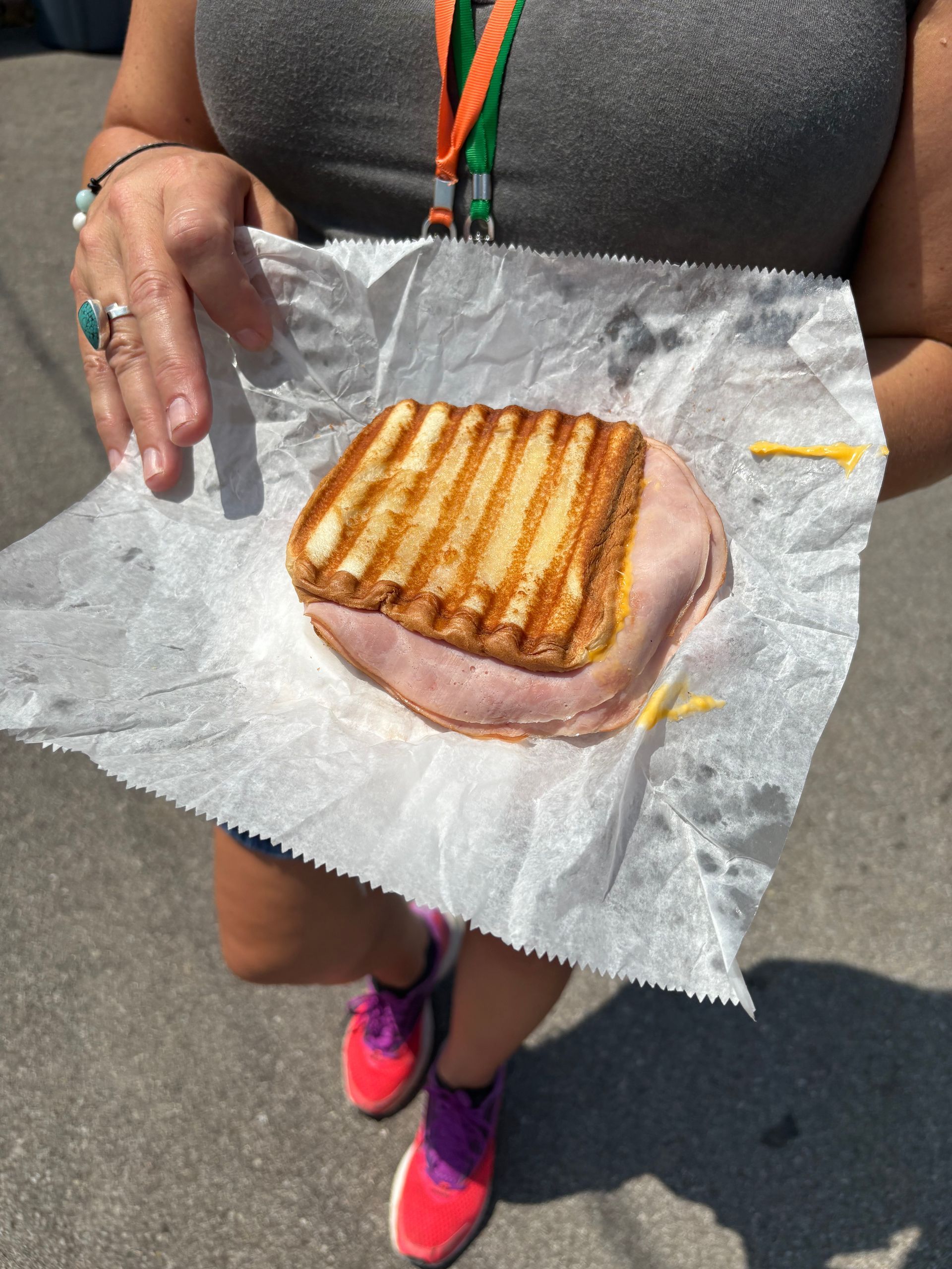 Person holding a grilled sandwich on wax paper outdoors, wearing a gray top and pink shoes.