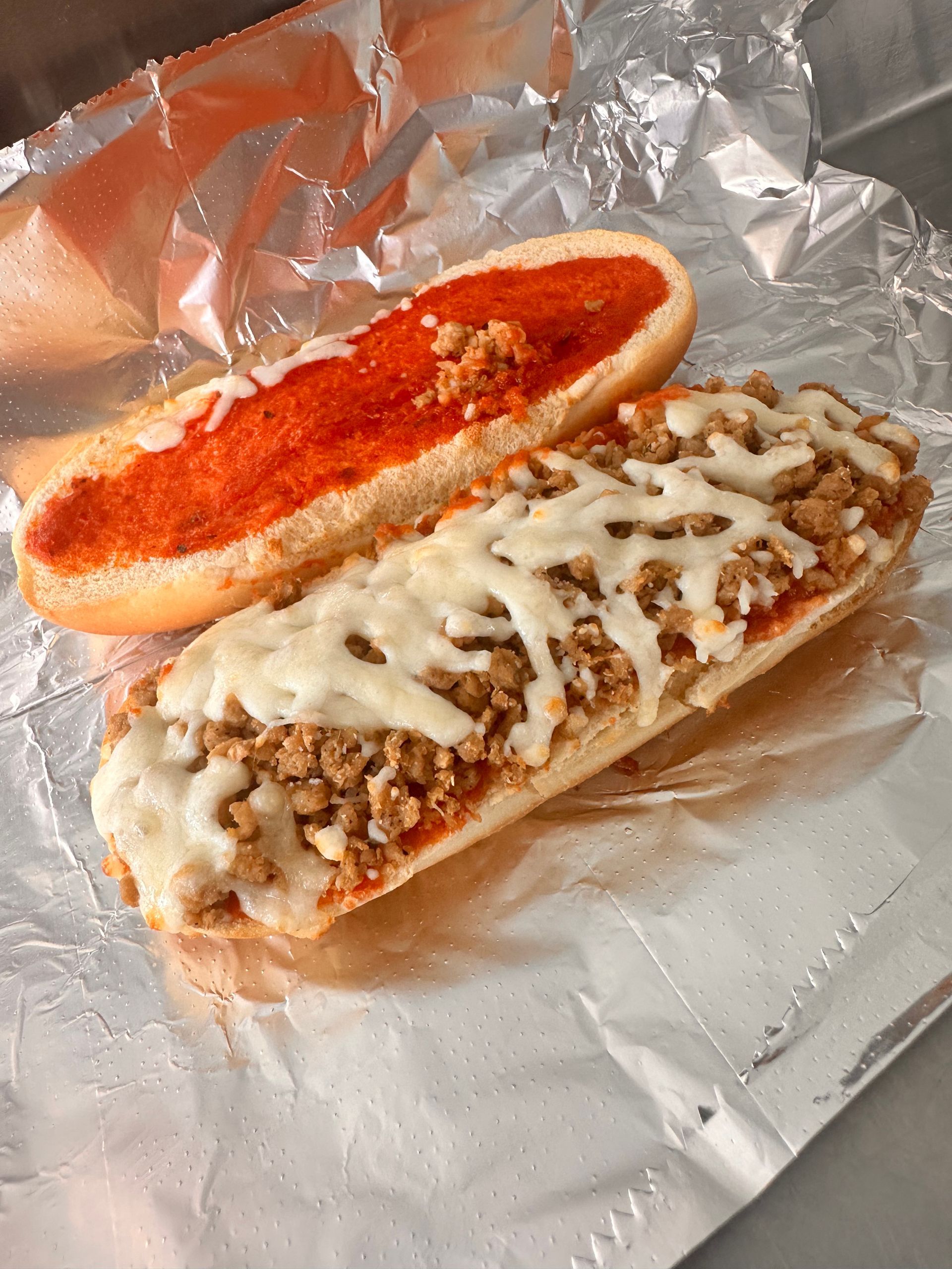 Two hot dog buns on foil, one with red sauce and one topped with meat and melted cheese.