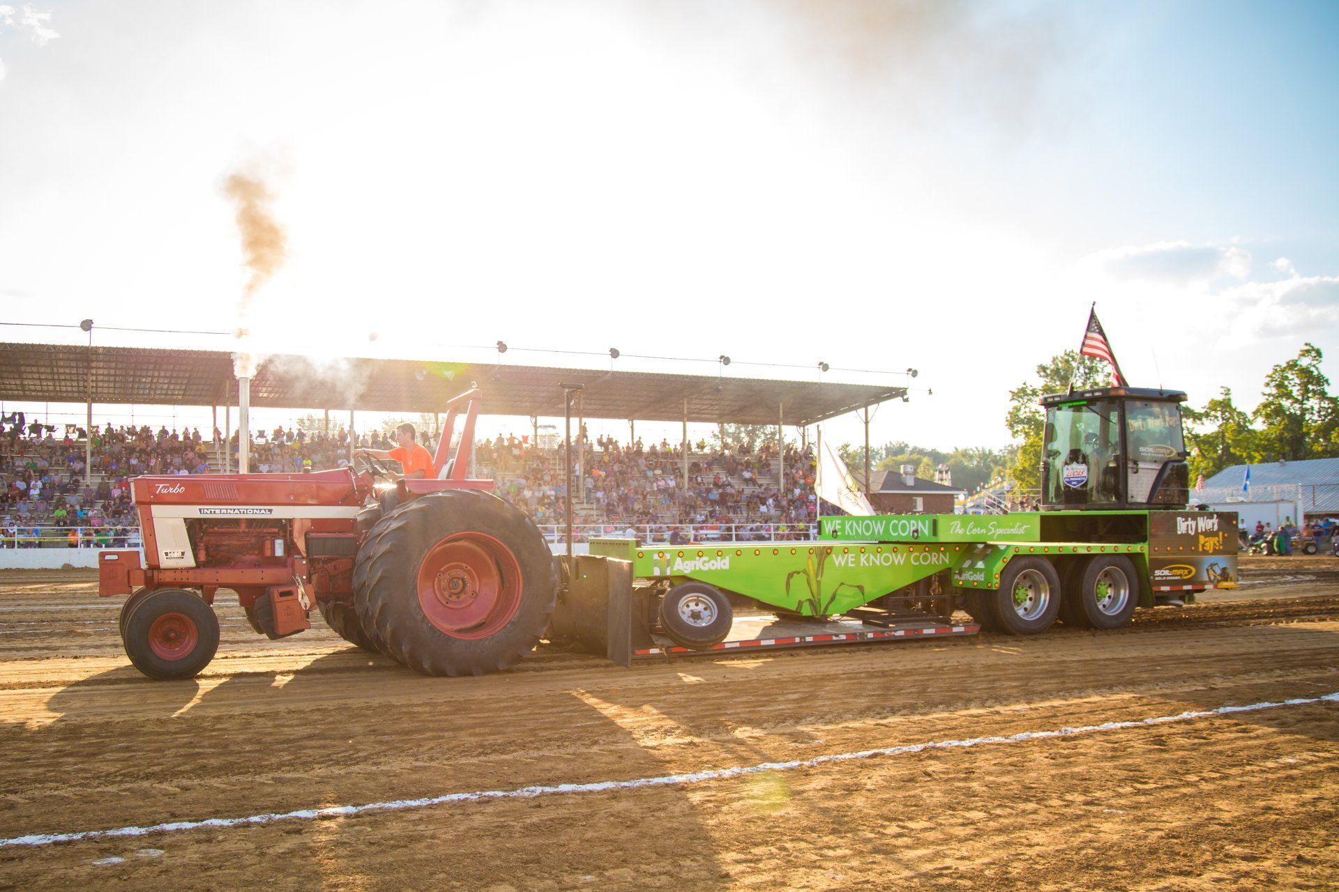 Red tractor pulling a green sled in a dirt arena with spectators in the background