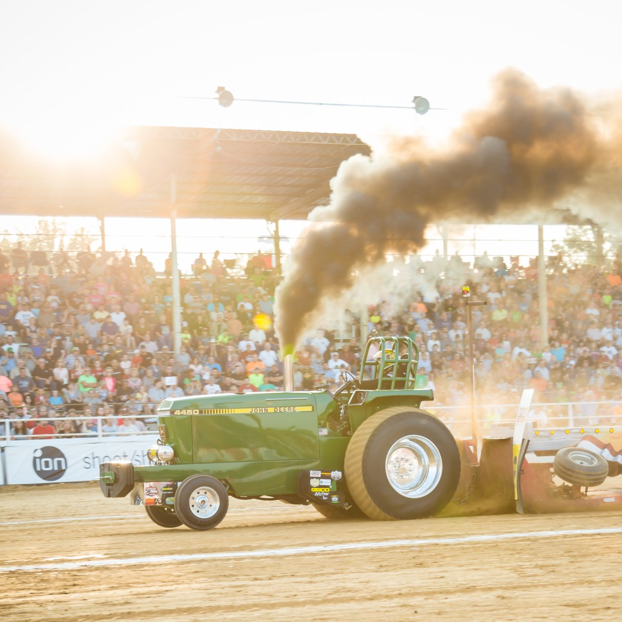 Green tractor pulling a sled, belching black smoke at a crowded dirt track event