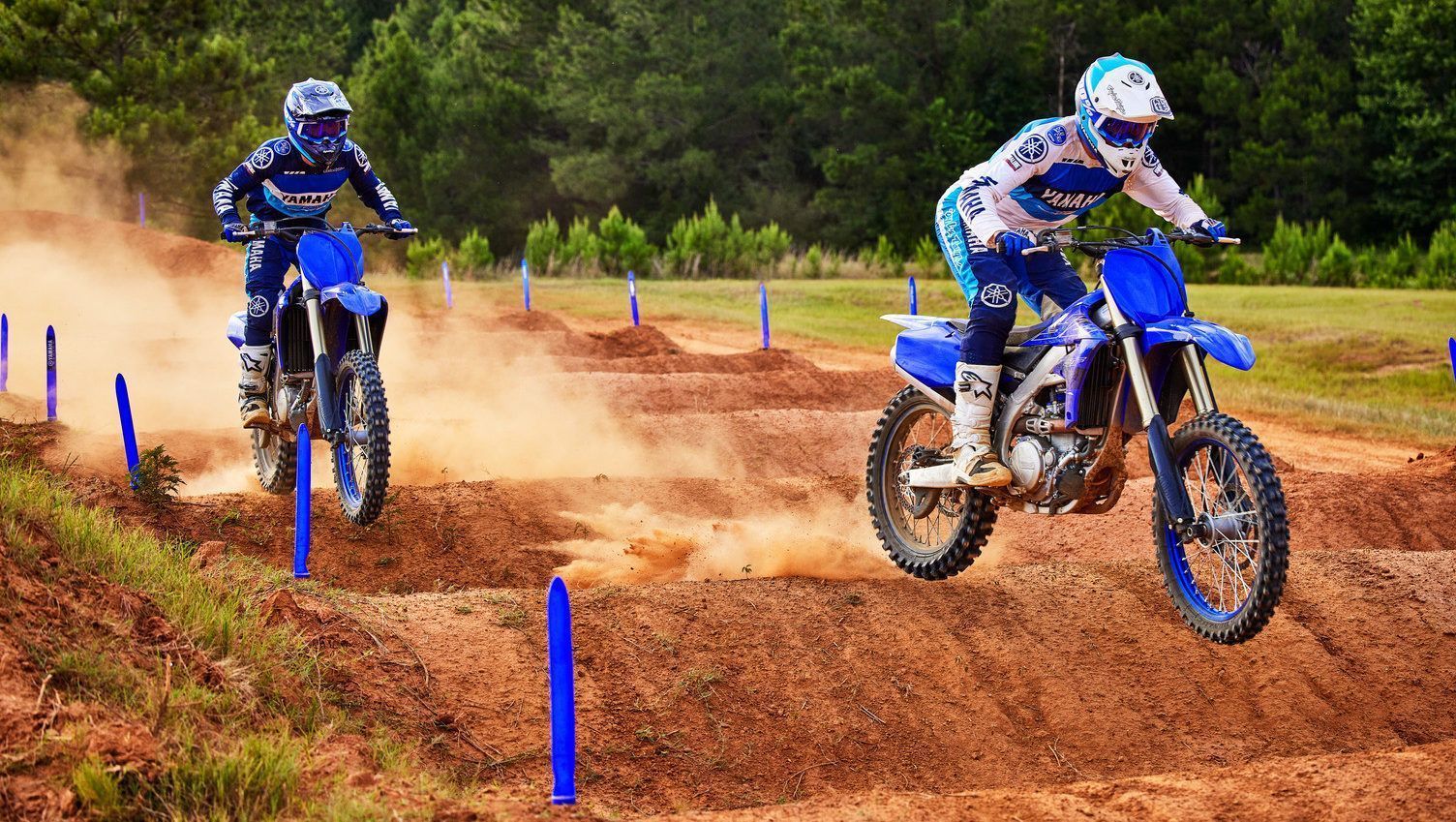 Two dirt bike riders race on a muddy track, kicking up dust near blue course markers.