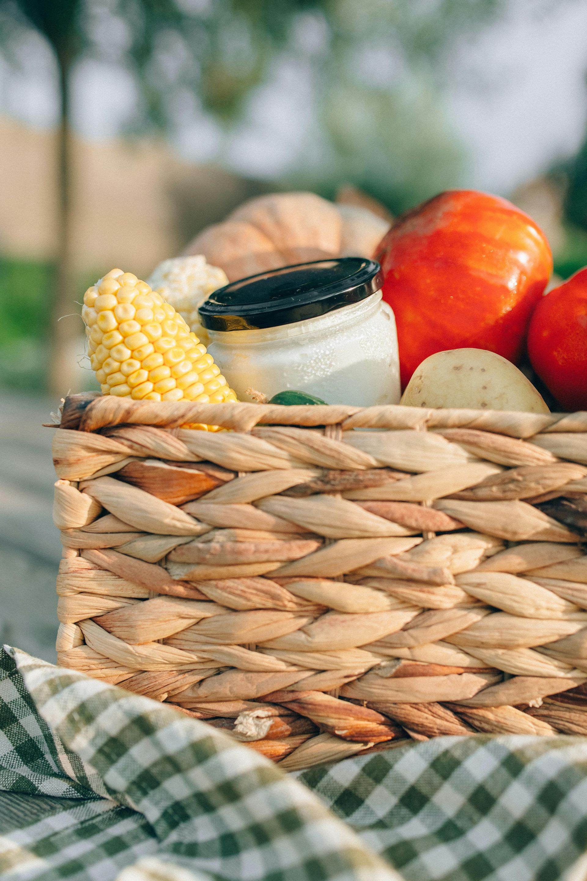 Woven basket of fresh corn, tomatoes, and jars on a green checkered cloth outdoors