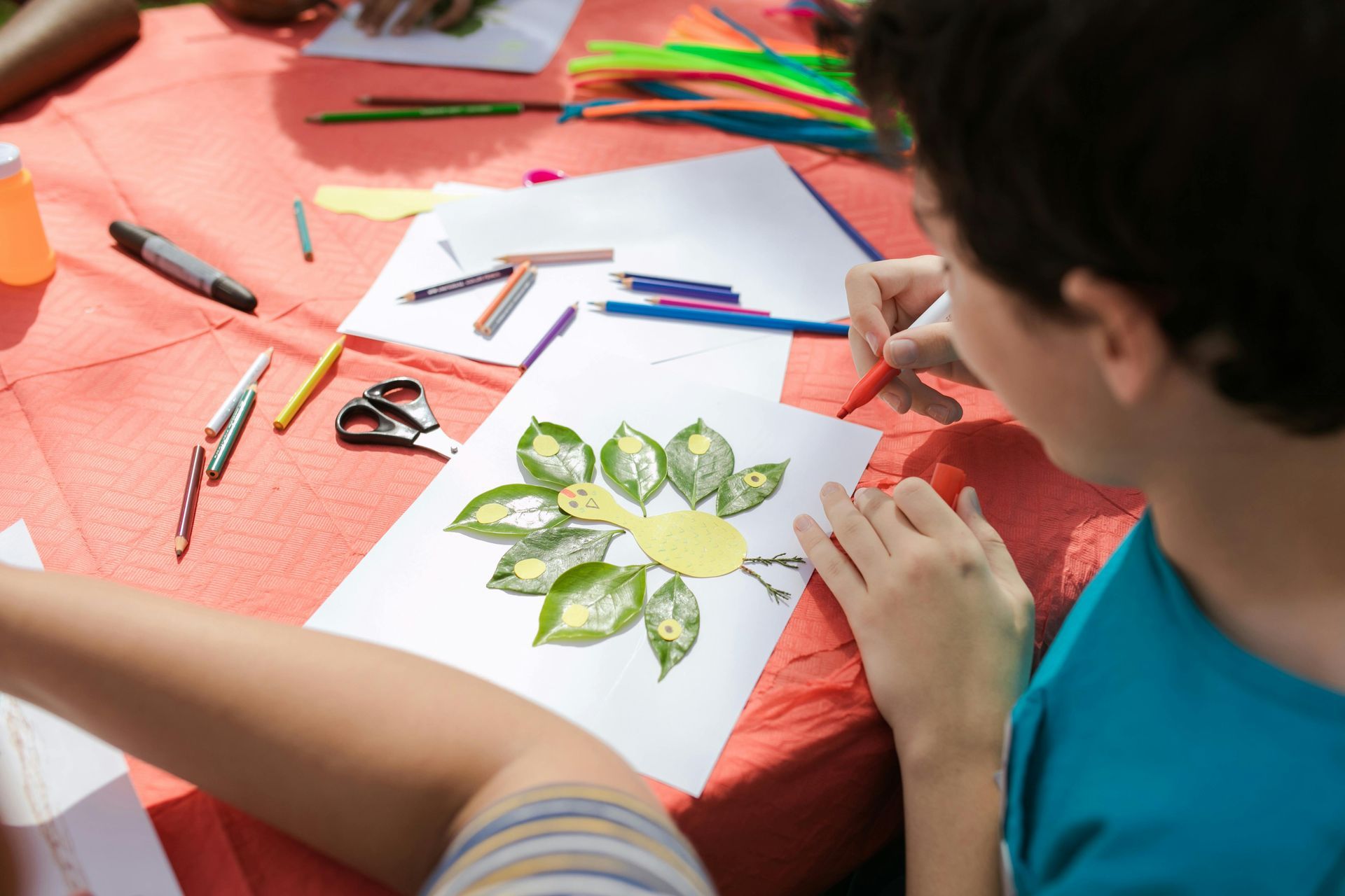 Person drawing a green flower design on white paper at a craft table with crayons and markers