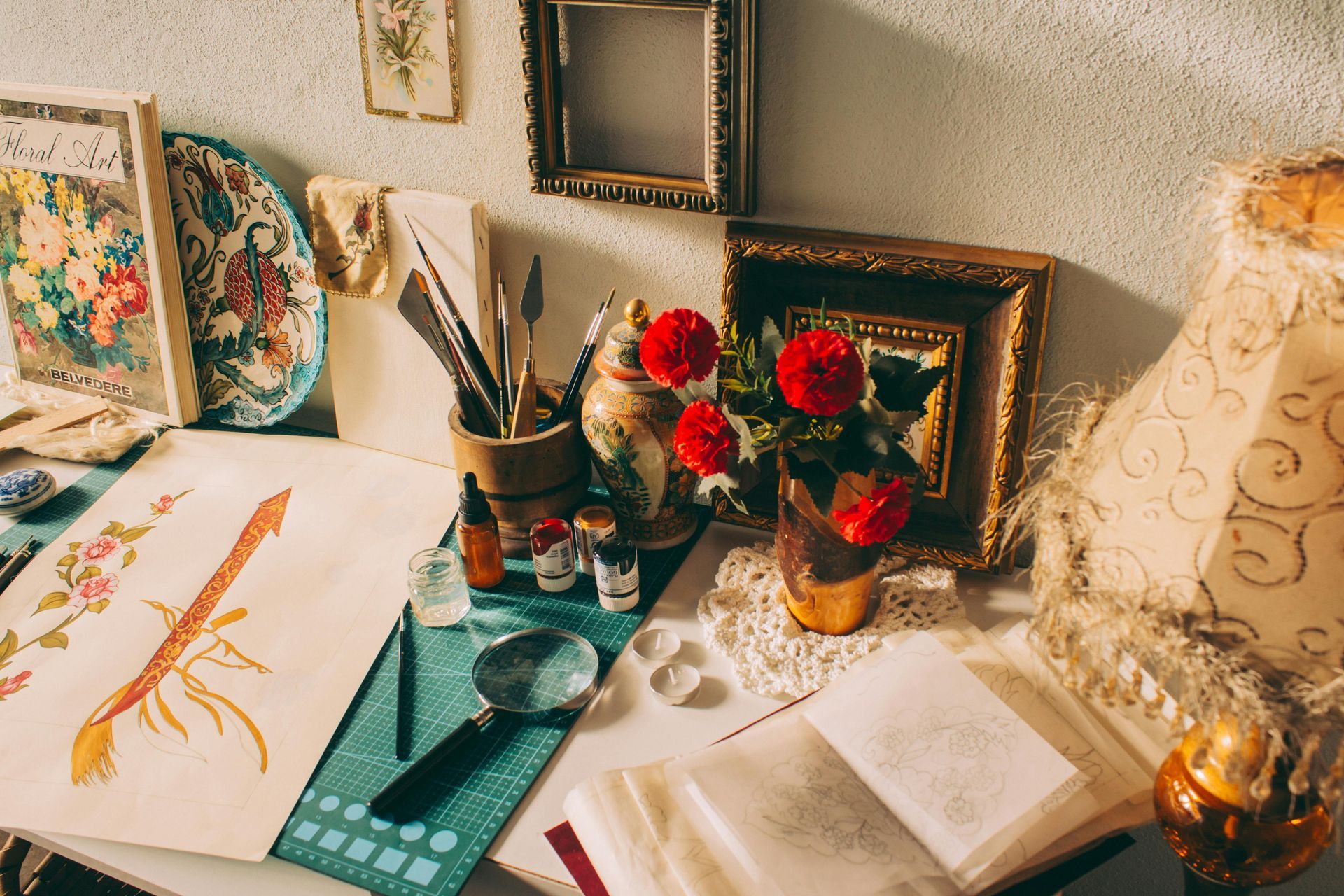 Art desk with sketchbook, paint supplies, flowers, and a lamp by a wall of framed art