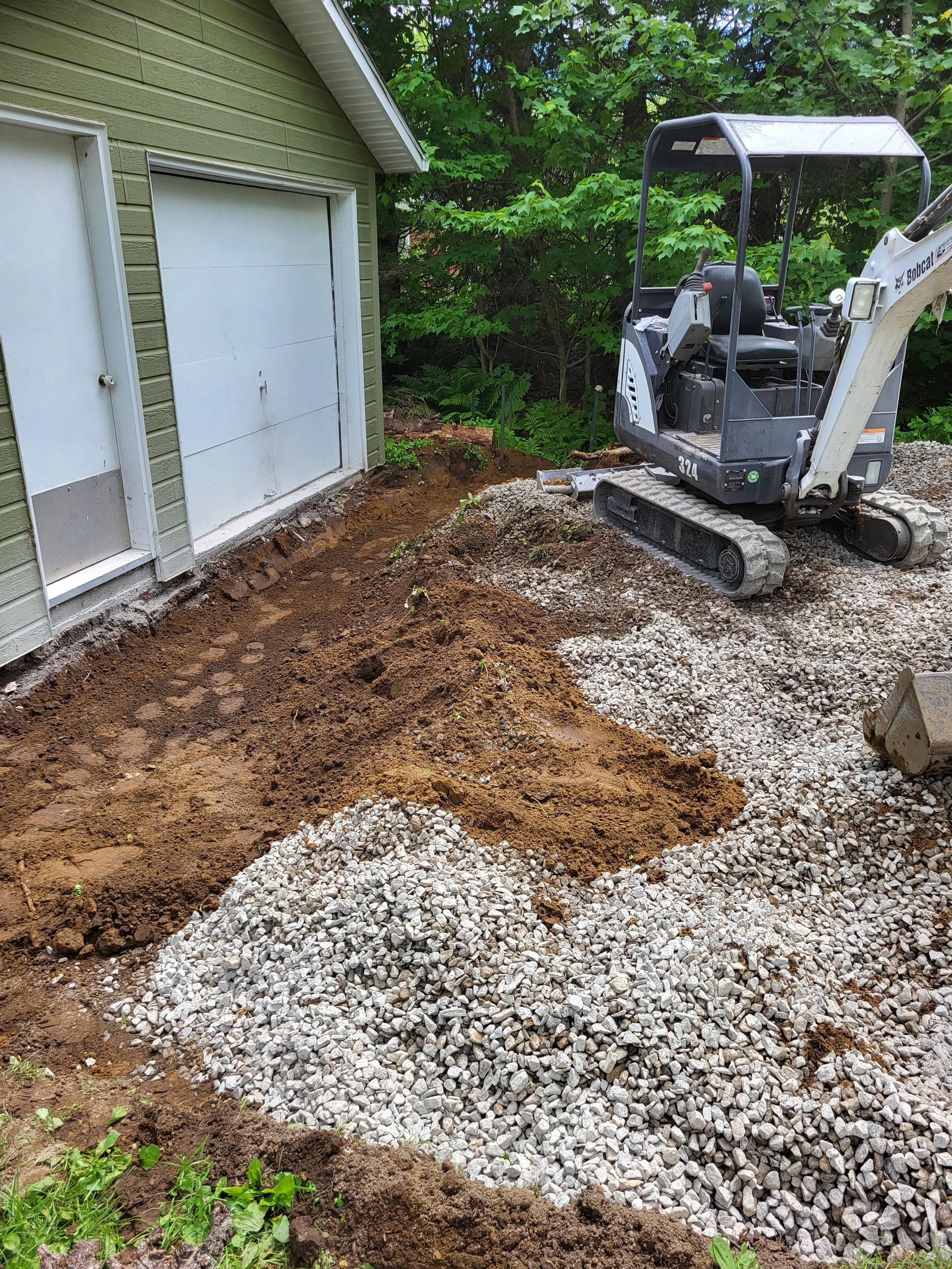 Une petite excavatrice est posée sur un tas de gravier devant un garage.