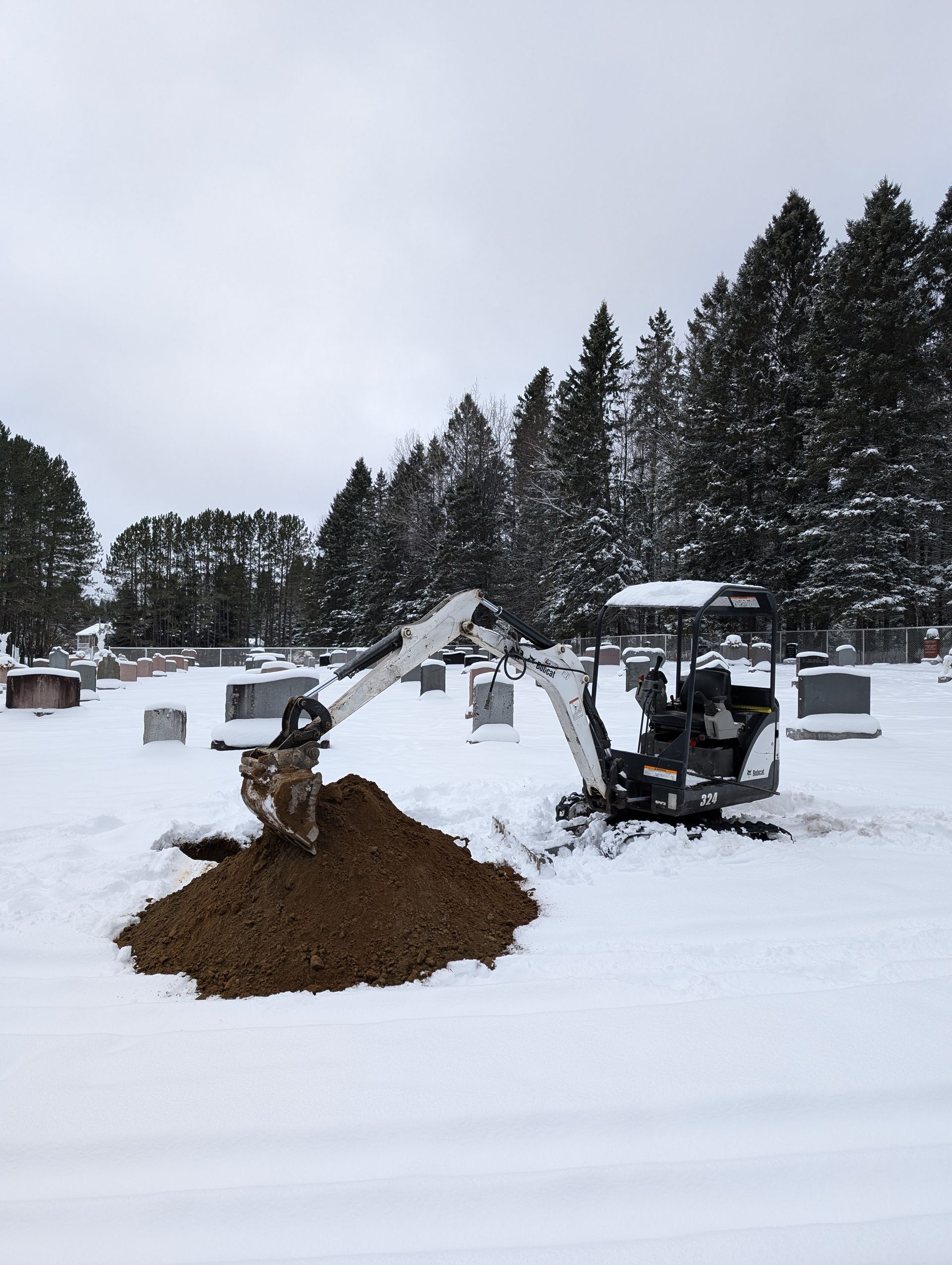 Une excavatrice creuse un trou dans la neige dans un cimetière.