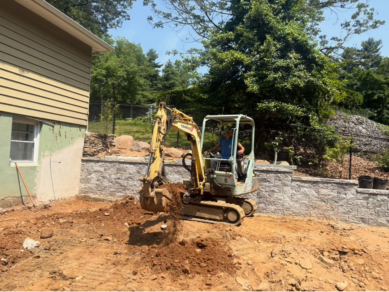 A man is driving an excavator in a dirt field in front of a house.