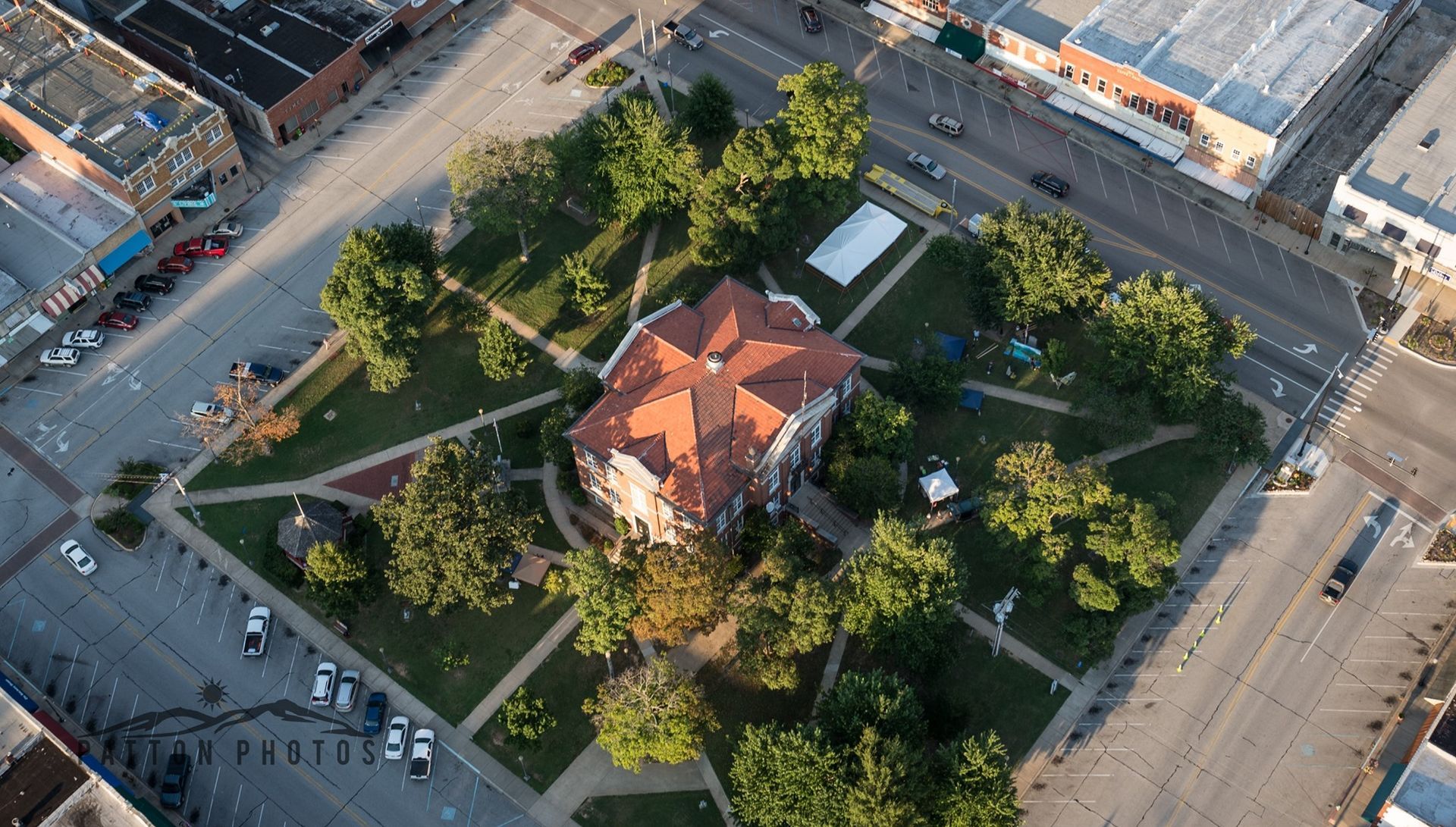 Aerial view of a red-roofed building surrounded by green grass and trees in a square park, with streets and buildings.