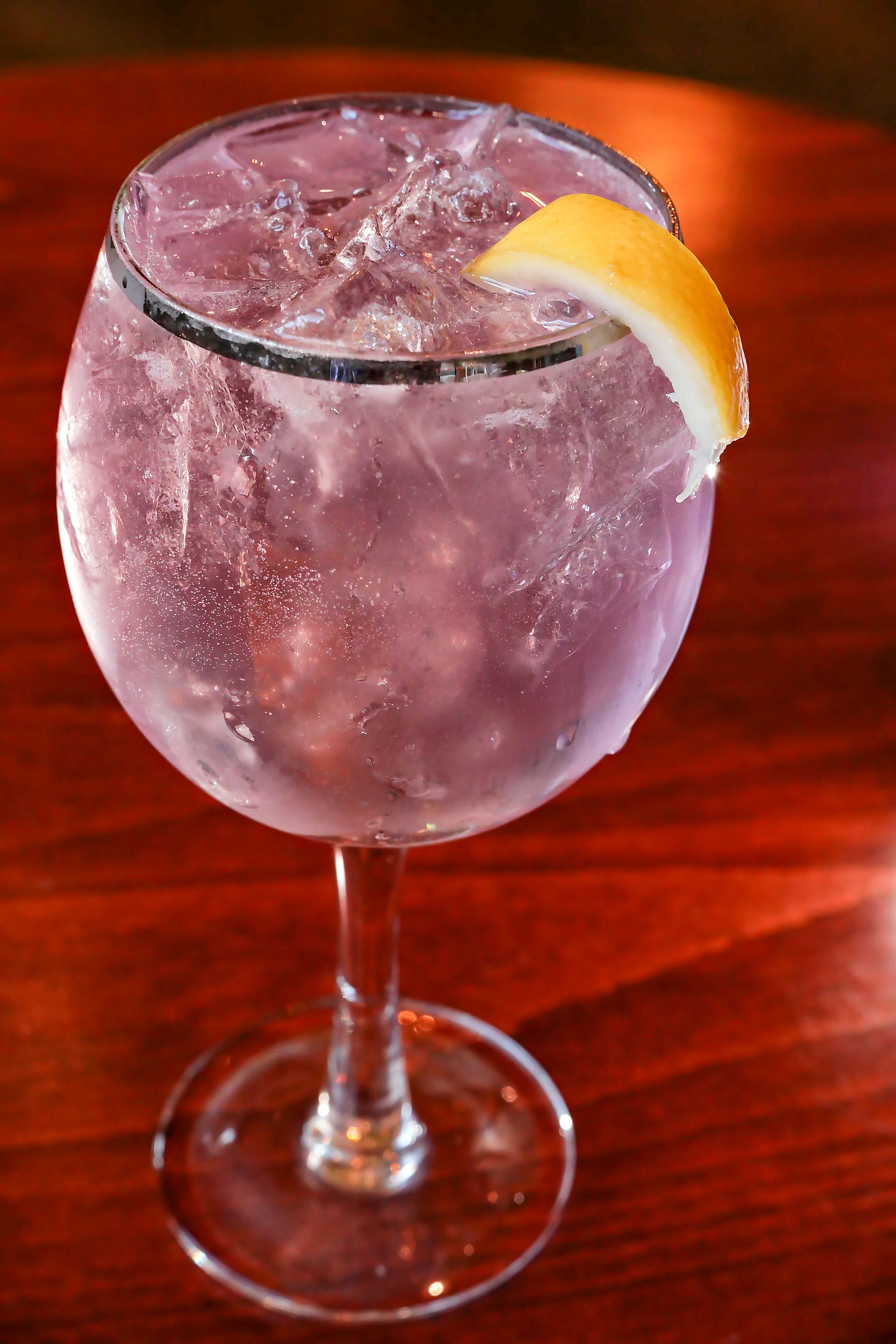 Purple cocktail in a large glass with lemon garnish, on a dark wooden table.