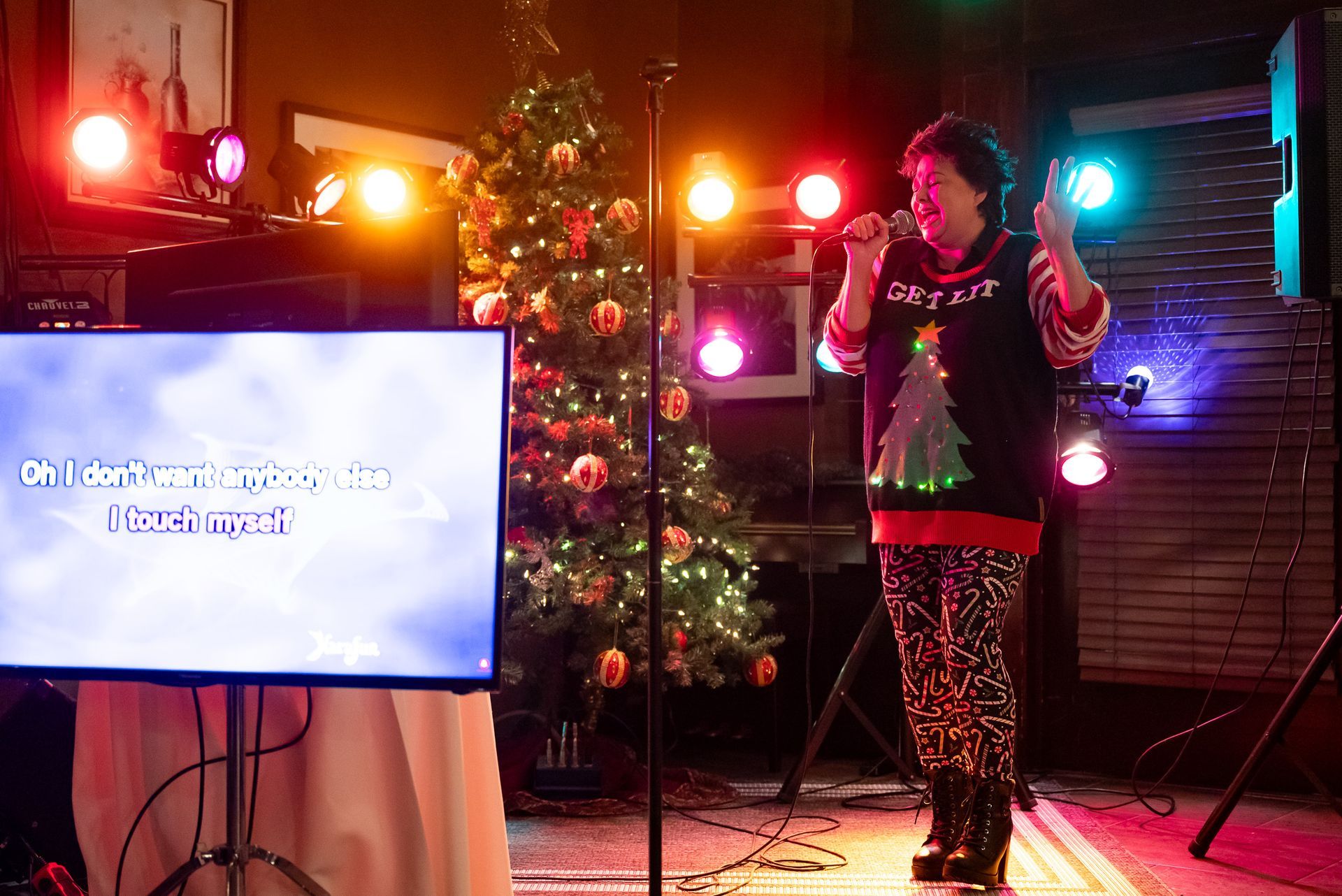 Woman singing karaoke at Christmas, wearing festive sweater and pants. Stage with tree and lights.