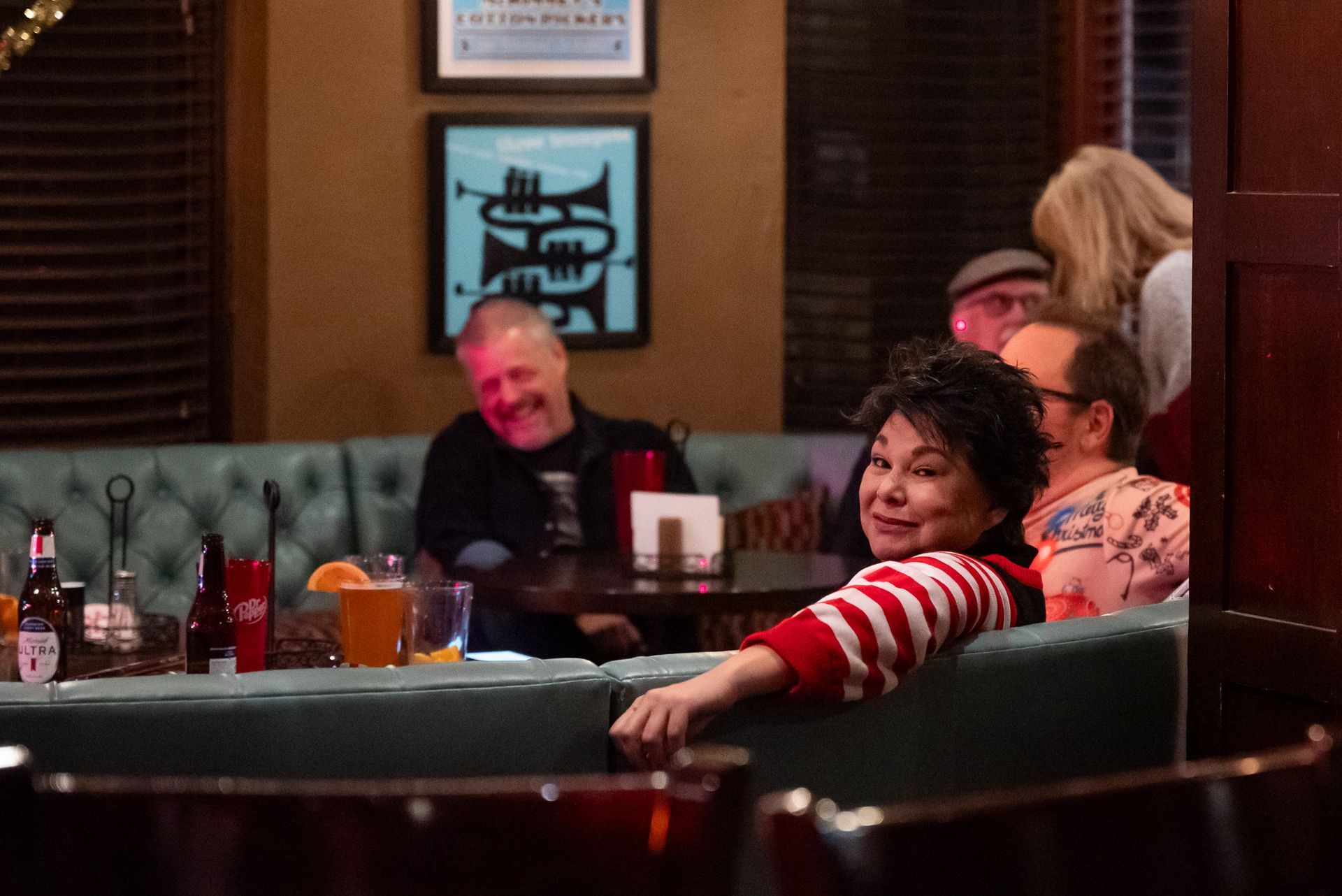 People at a bar, smiling and seated at a booth. Dark interior with artwork on the wall.