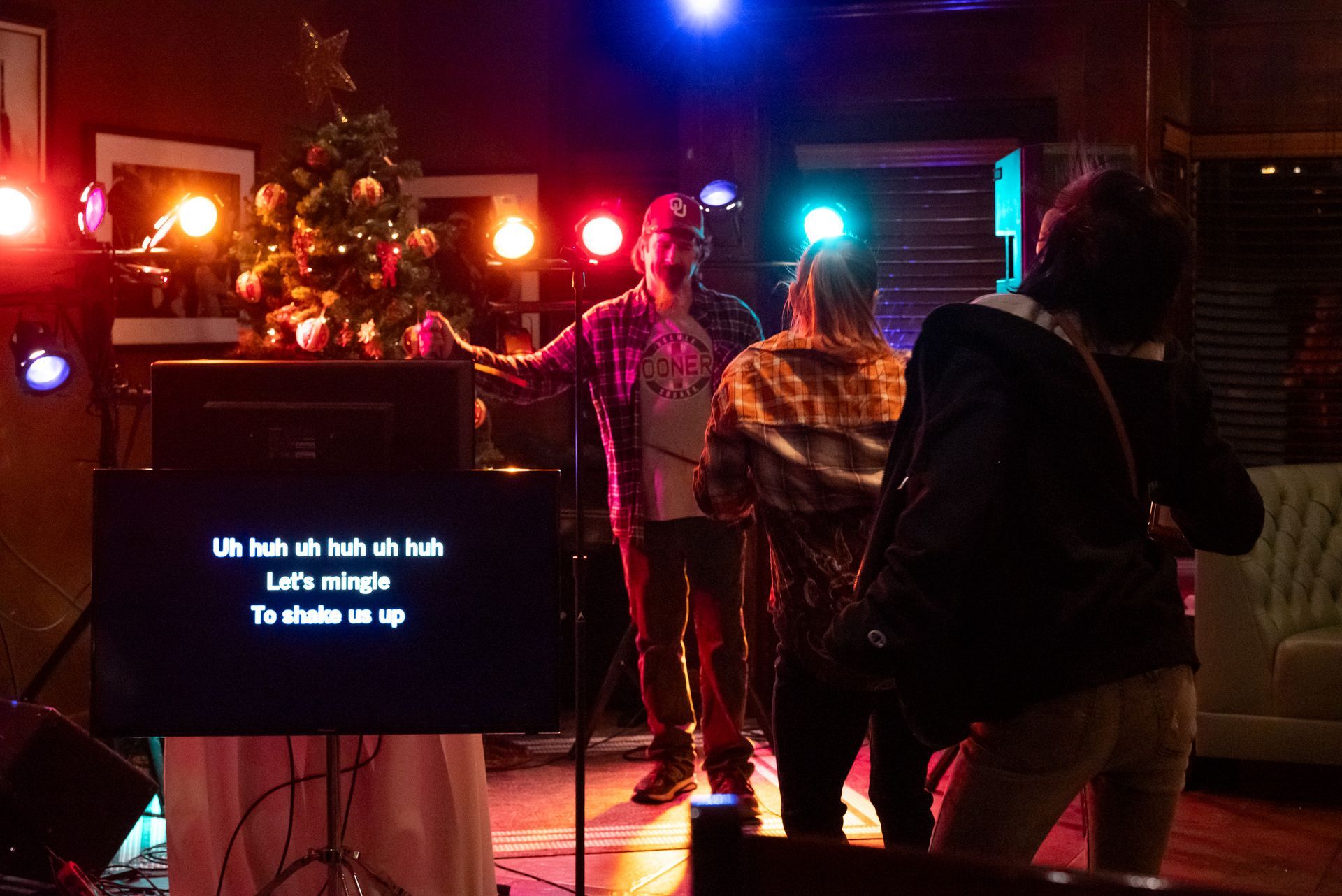 People sing karaoke in a dimly lit bar. A screen displays lyrics. A Christmas tree is in the background.
