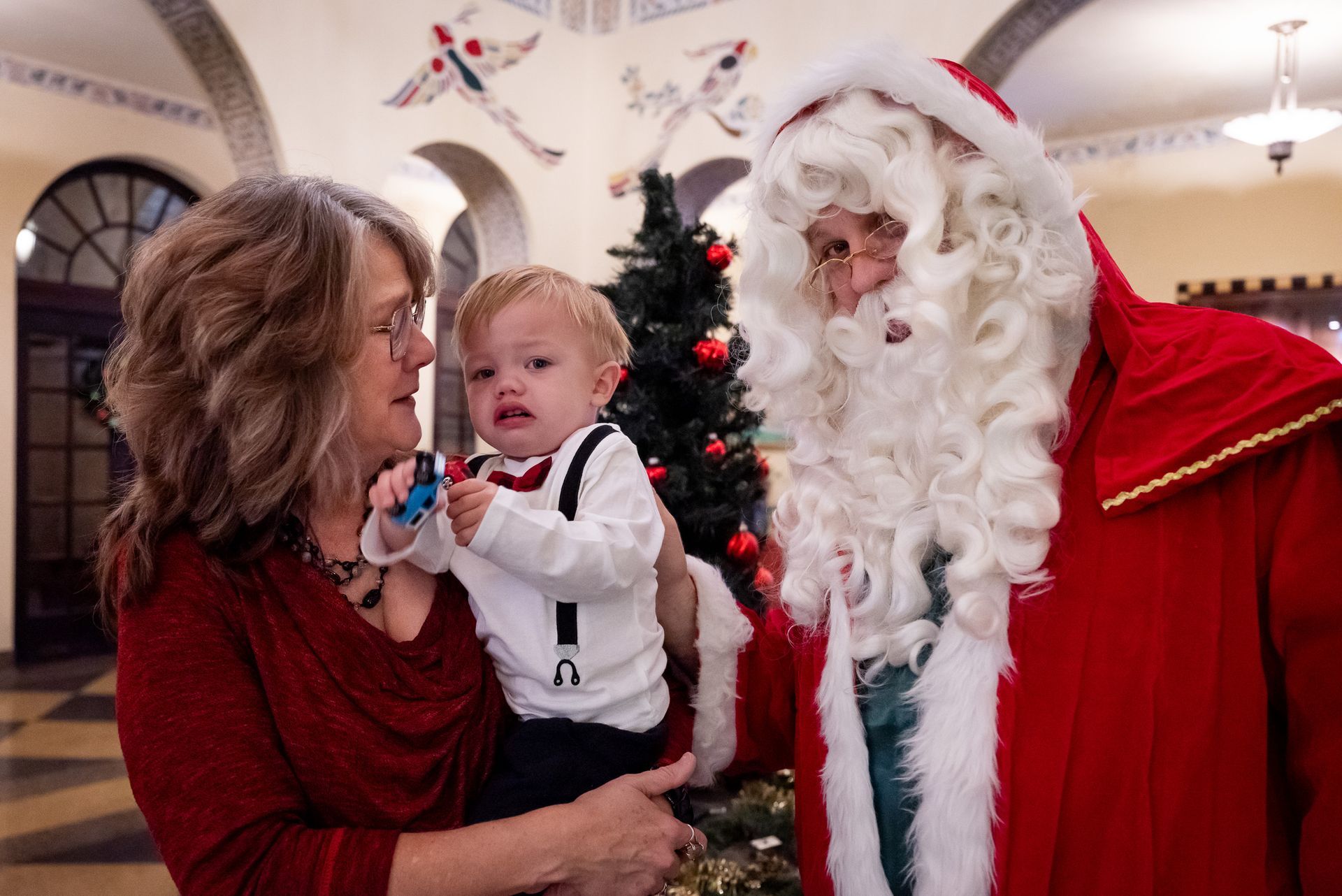 Woman holds crying baby near Santa Claus, festive setting, Christmas tree.