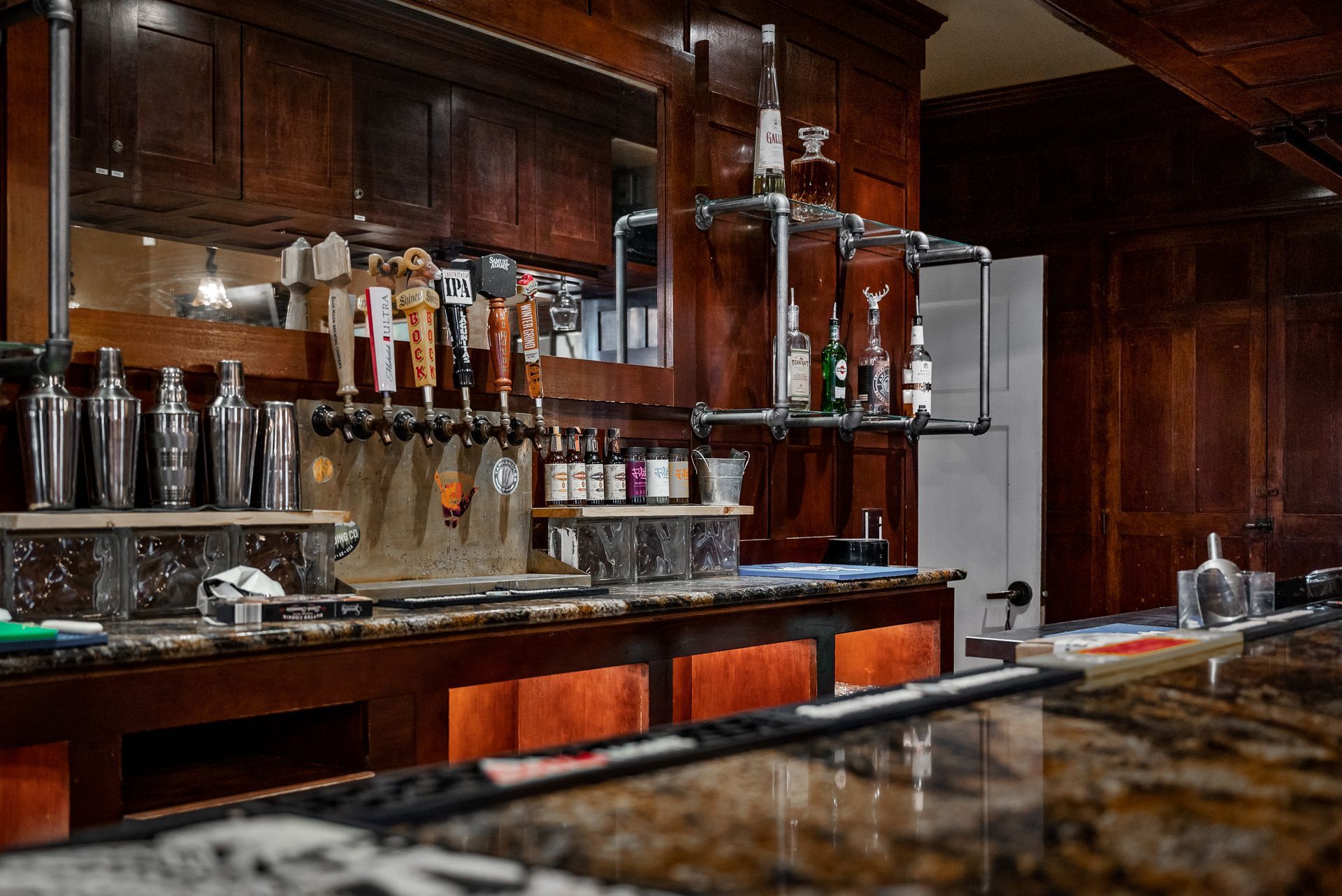 A bar with dark wood and granite countertop, beer taps, and bottles of liquor.