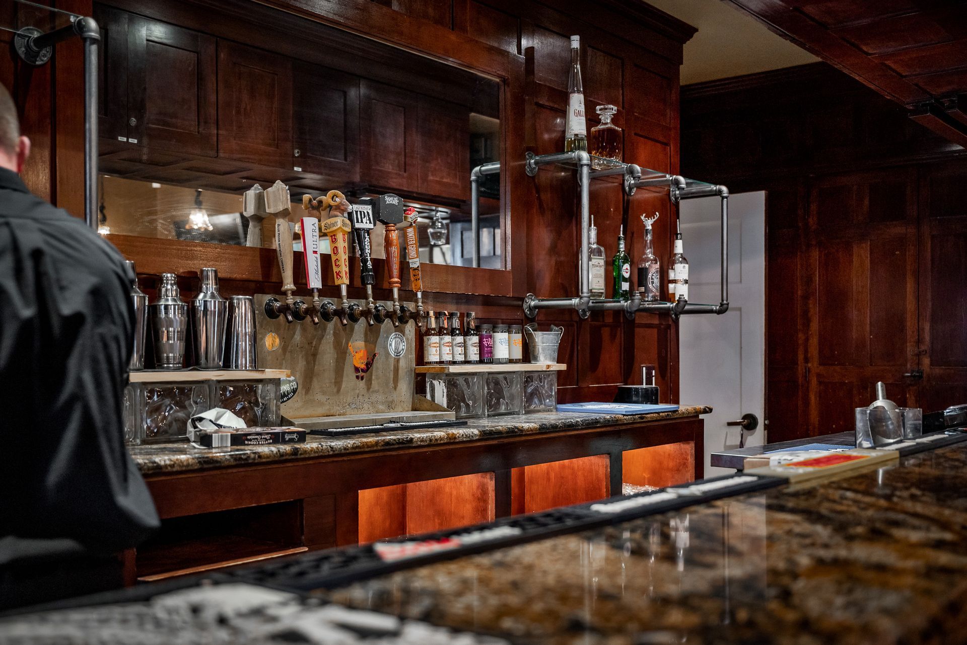 A dark, upscale bar with bartender, beer taps, liquor bottles, and a wood and stone countertop.
