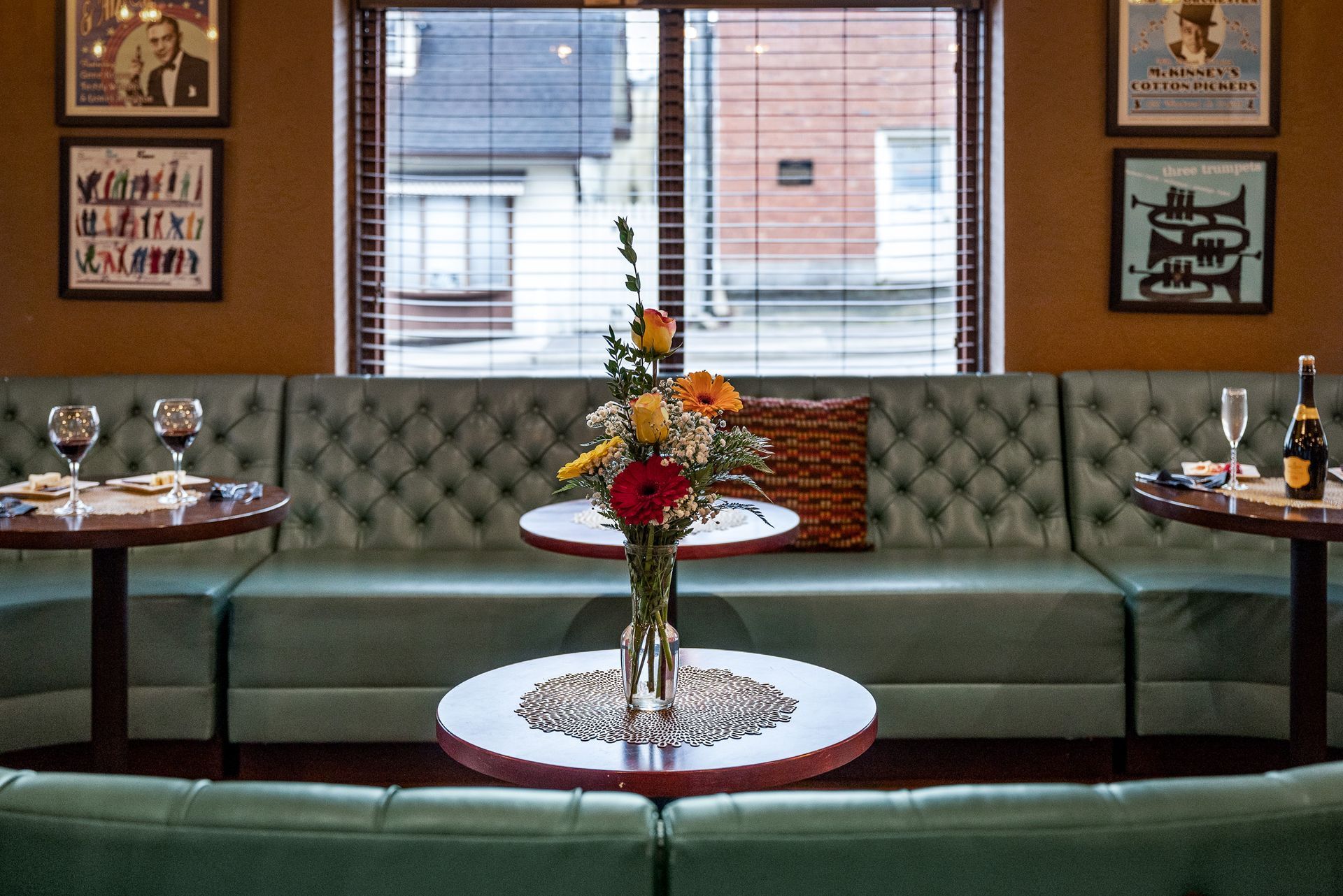 A restaurant booth with green seating, small tables, floral centerpiece, posters, and a window.
