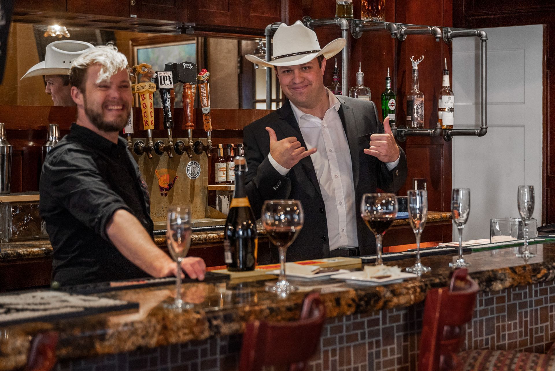 Two men in cowboy hats at a bar; one has foam on his head and is smiling, other makes a 