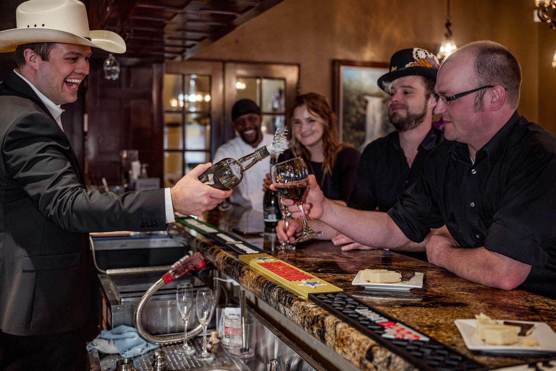 Bartender in cowboy hat pours drinks for customers at a bar; people smiling, celebrating, indoors.
