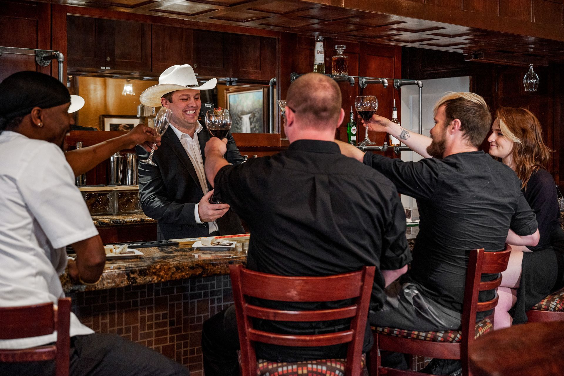 People toasting drinks at a bar; man in cowboy hat smiles.