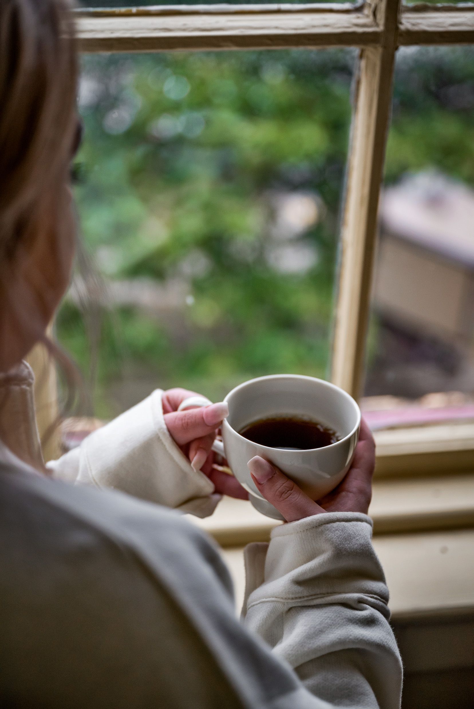 Woman holding coffee, looking out a window at a green, blurry background.